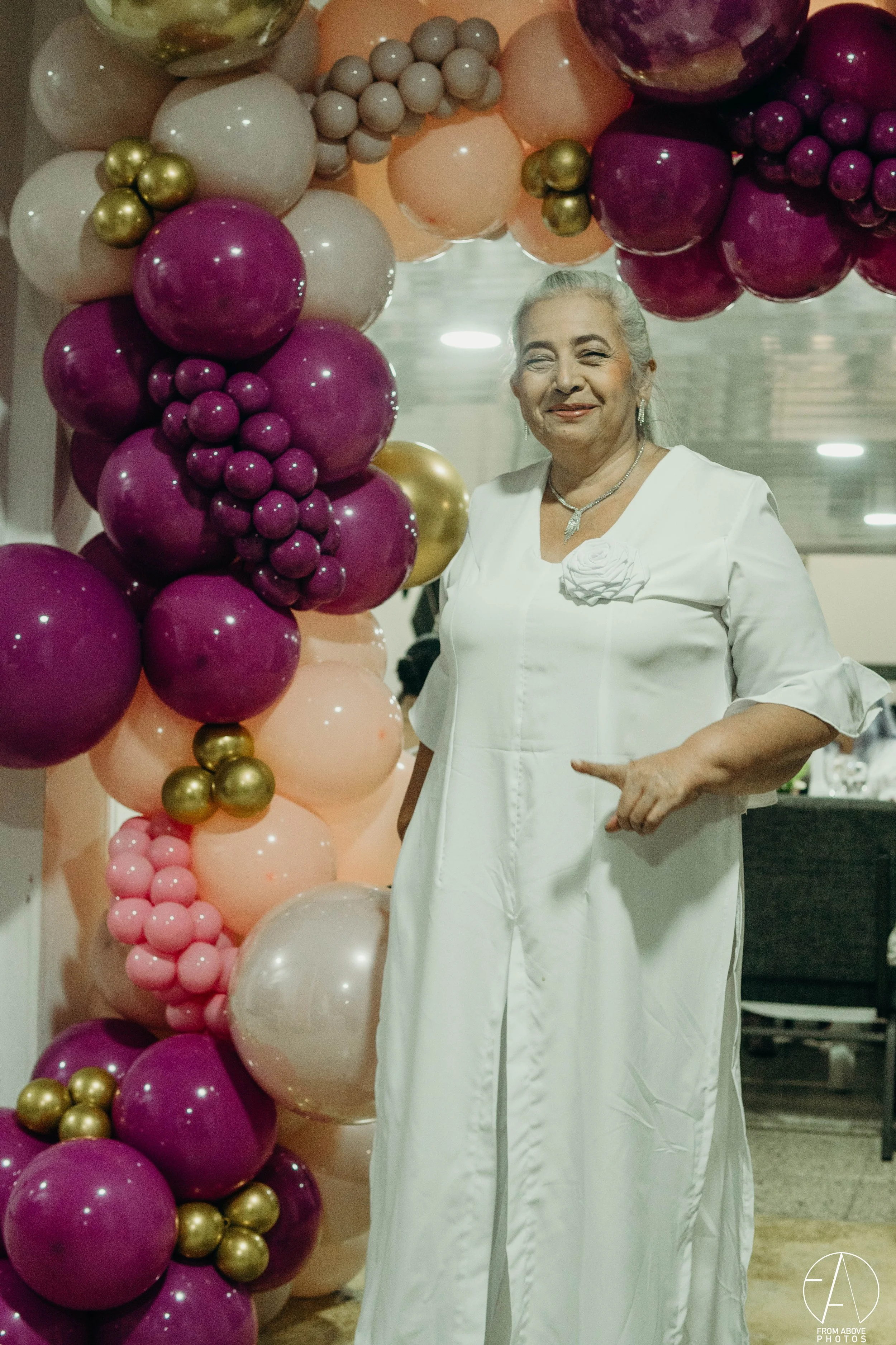 Anciana vestida de blanco sonriendo frente a un arco de globos de colores.