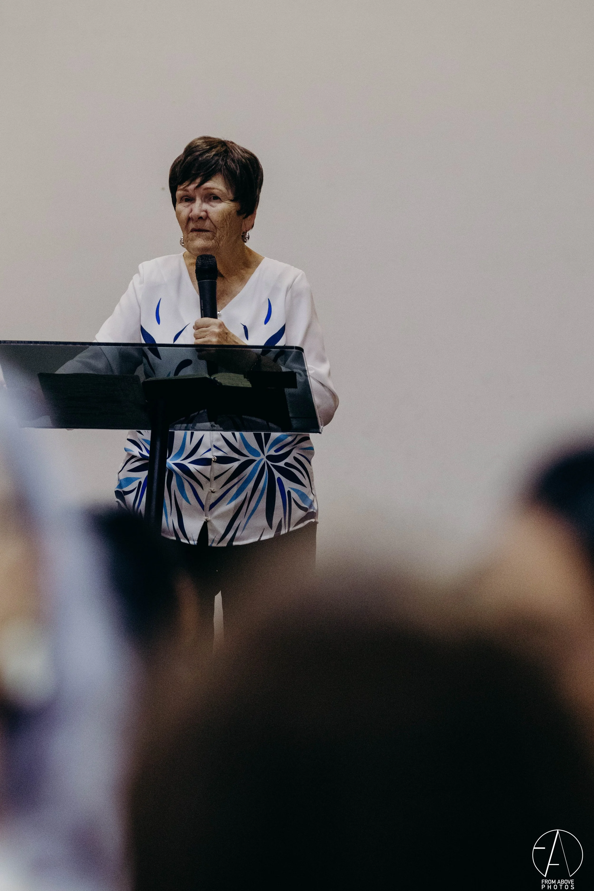 Una mujer de cabello corto y oscuro, vestida con blusa blanca y pantalones con un diseño azul y blanco, sostiene un micrófono mientras da un discurso frente a un fondo liso.