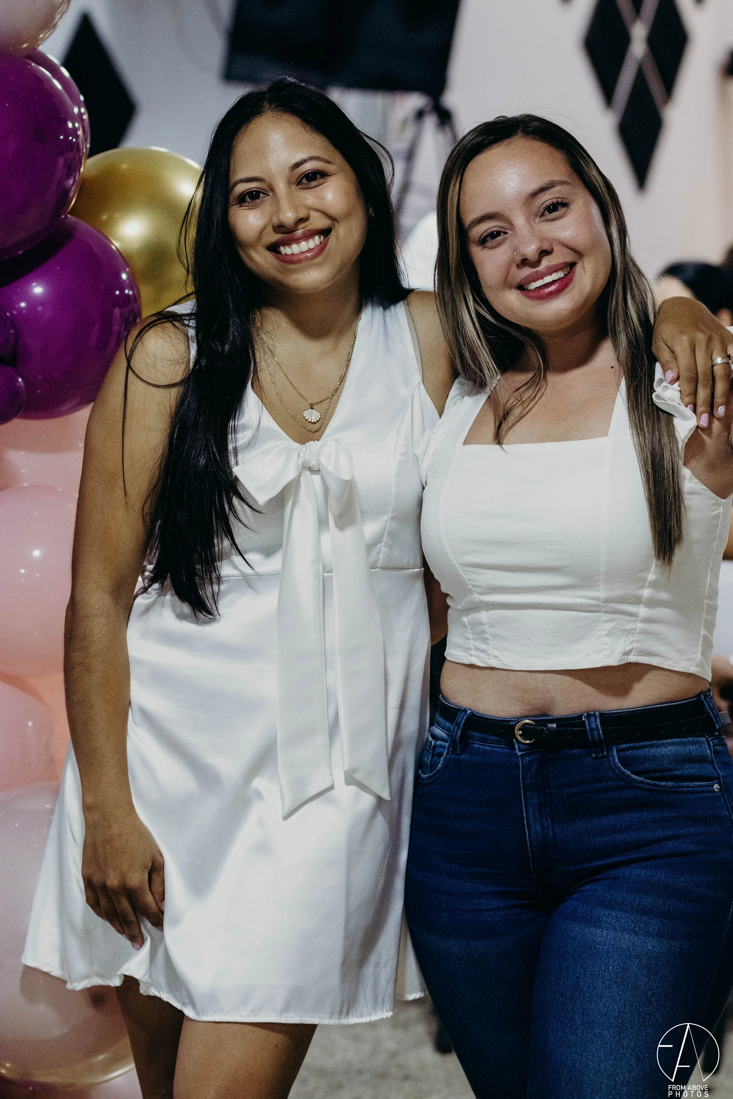 Dos mujeres sonriendo y posando juntas en una celebración, rodeadas de globos morados y dorados.