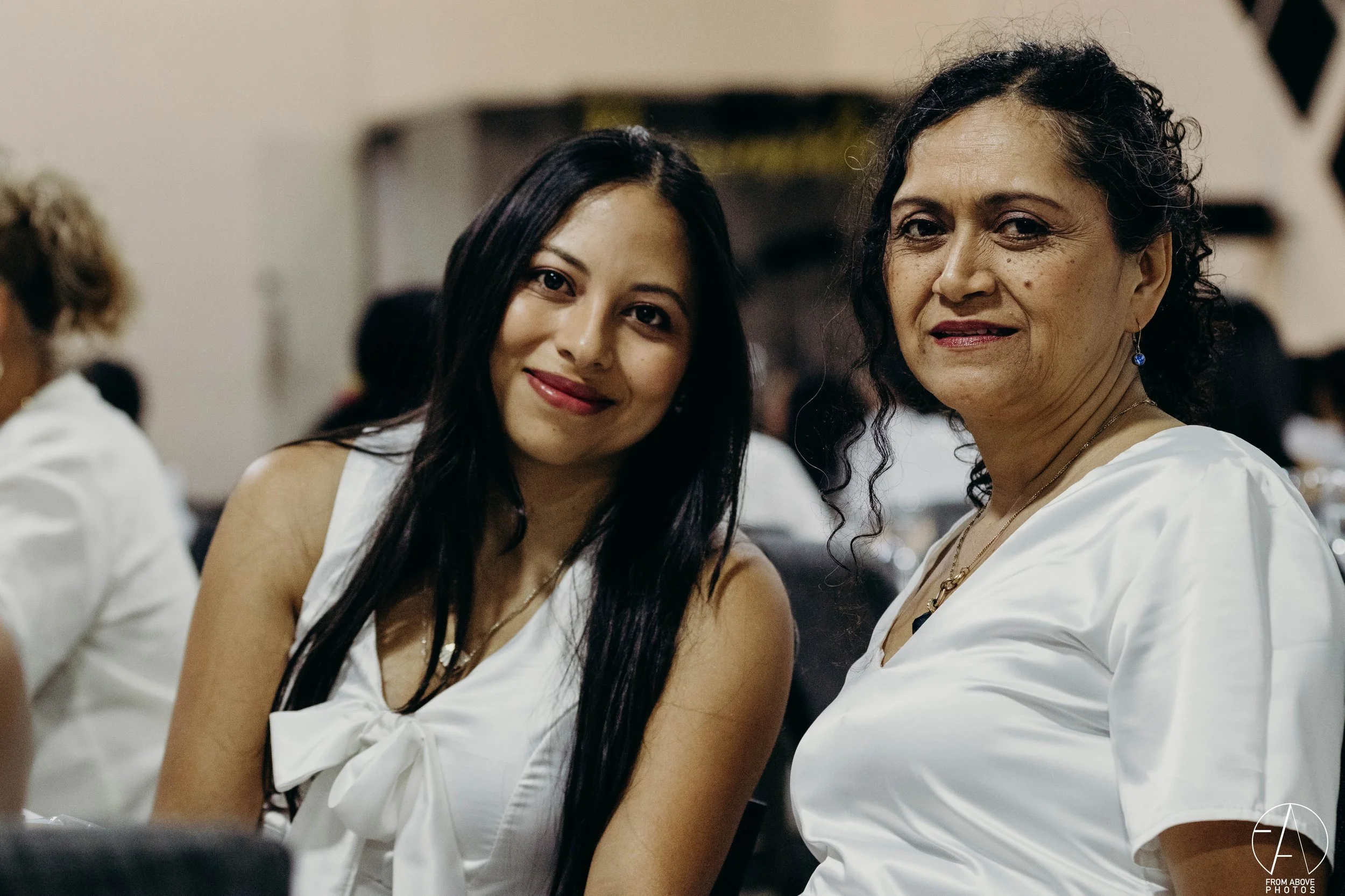 Dos mujeres sentadas en una reunión o evento formal, vestidas de blanco, con cabello largo y rizado, y pelo oscuro y liso, respectivamente.