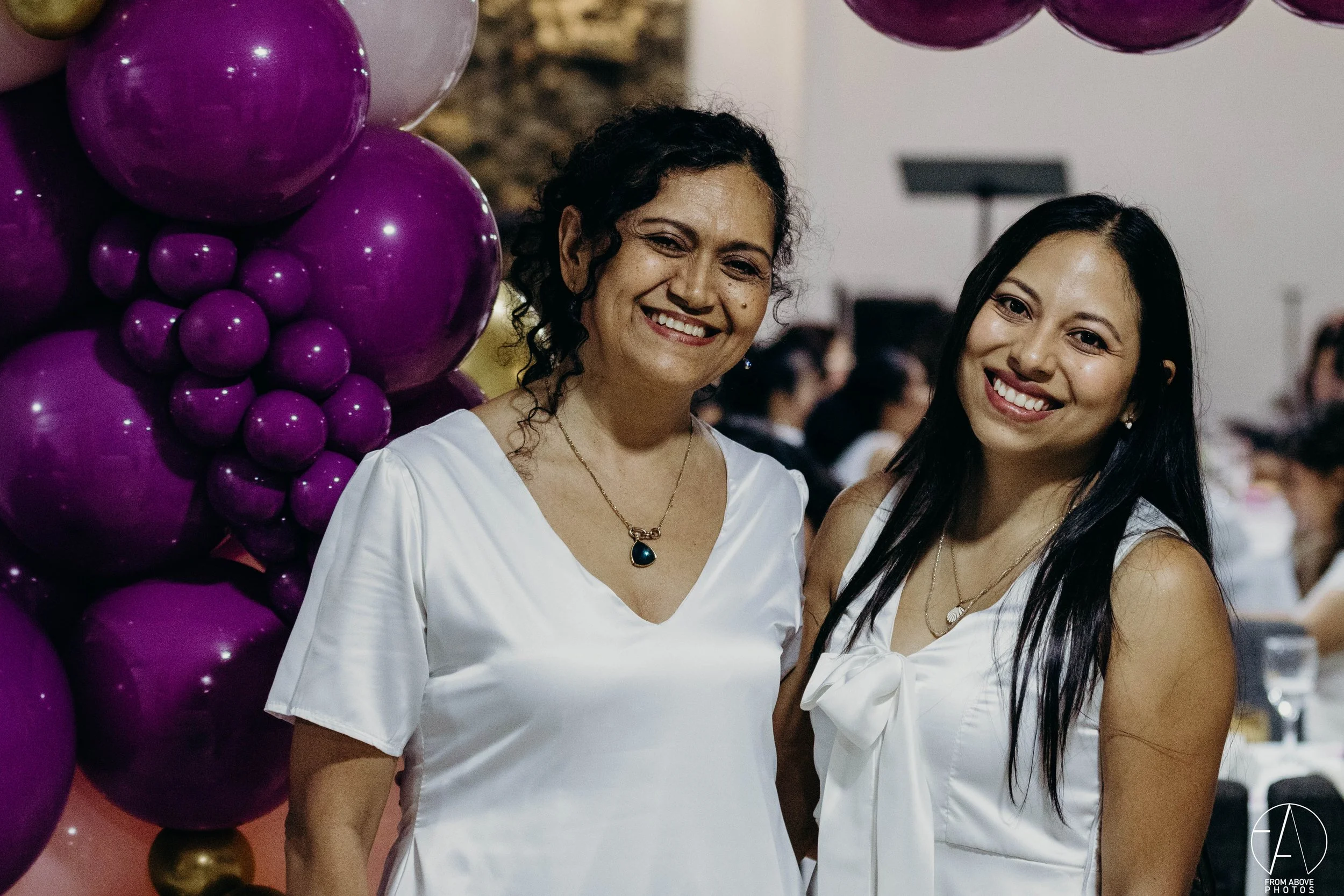 Dos mujeres jóvenes con vestidos blancos sonriendo en una celebración, con globos morados y burdeos en el fondo.