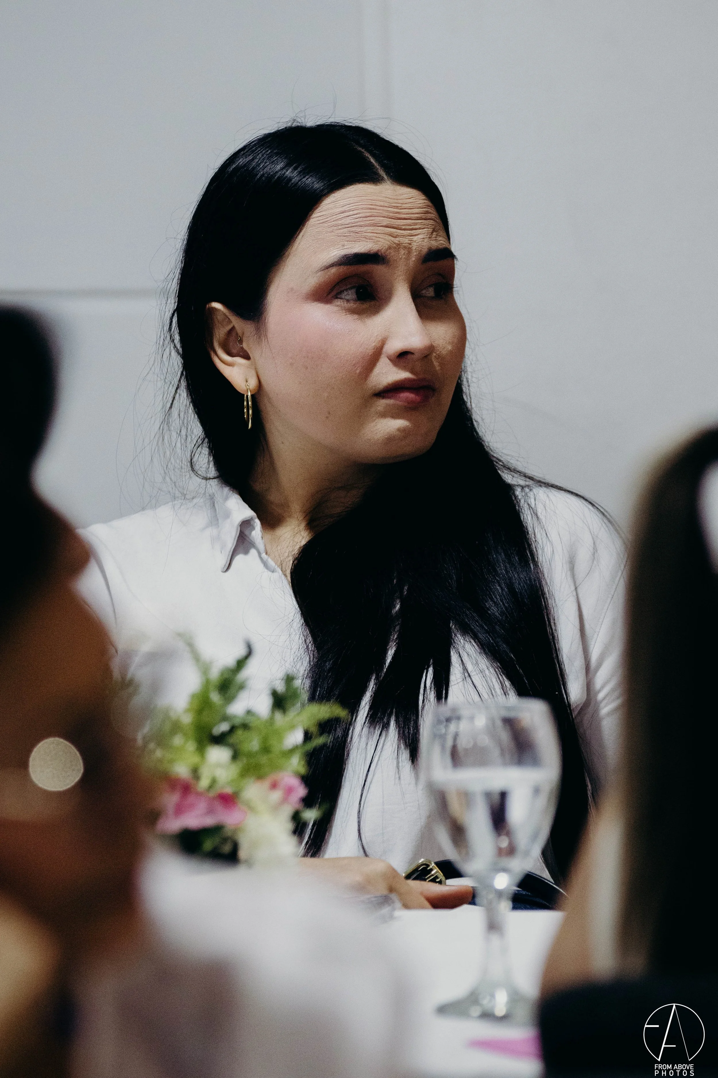 Mujer de cabello largo y oscuro, con orejas de aro, mirando a un lado en una reunión o evento social, con un vaso de agua y flores en la mesa.