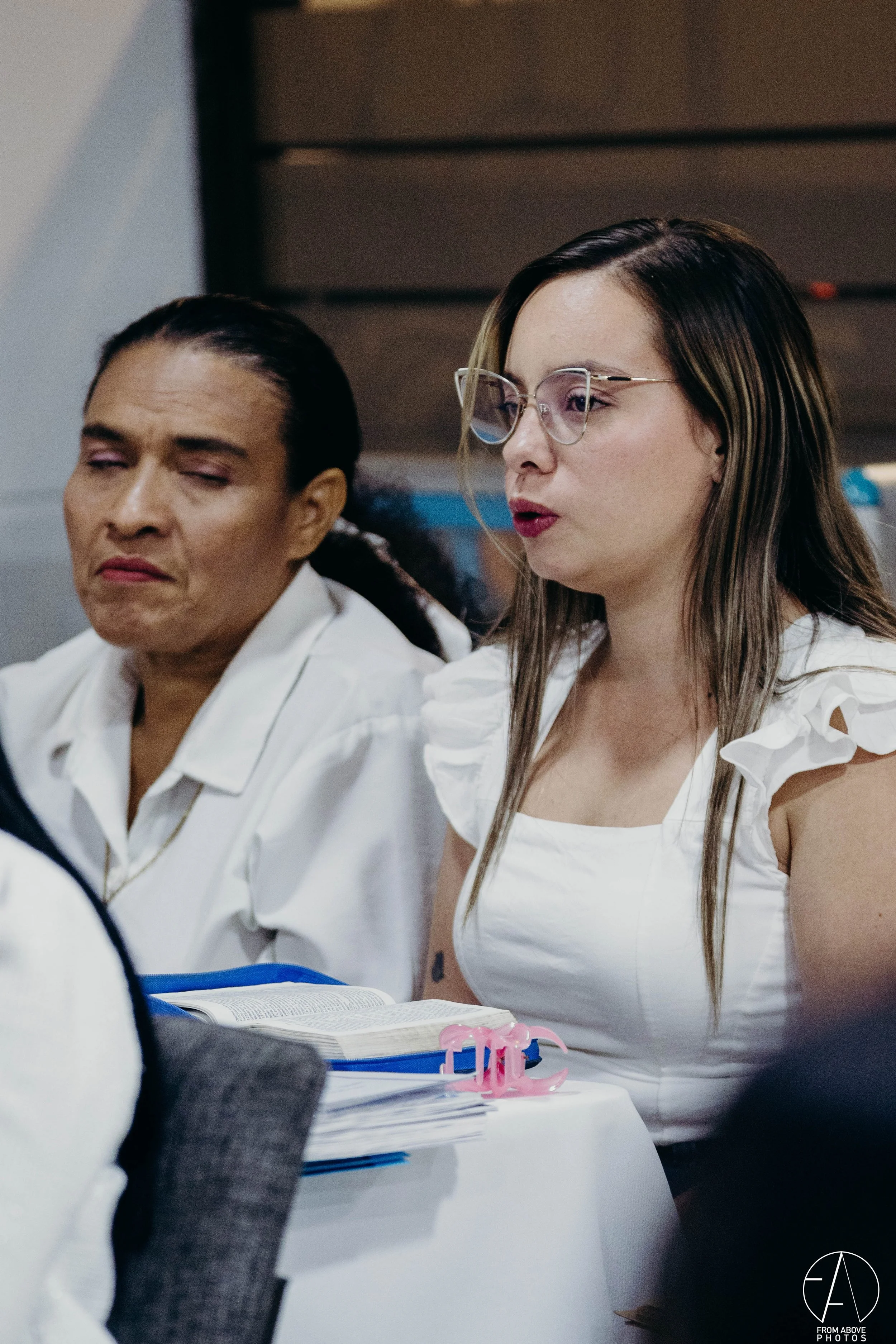 Dos mujeres sentadas en una mesa, una leyendo un libro abierto, con documentos y un portapapeles frente a ella.