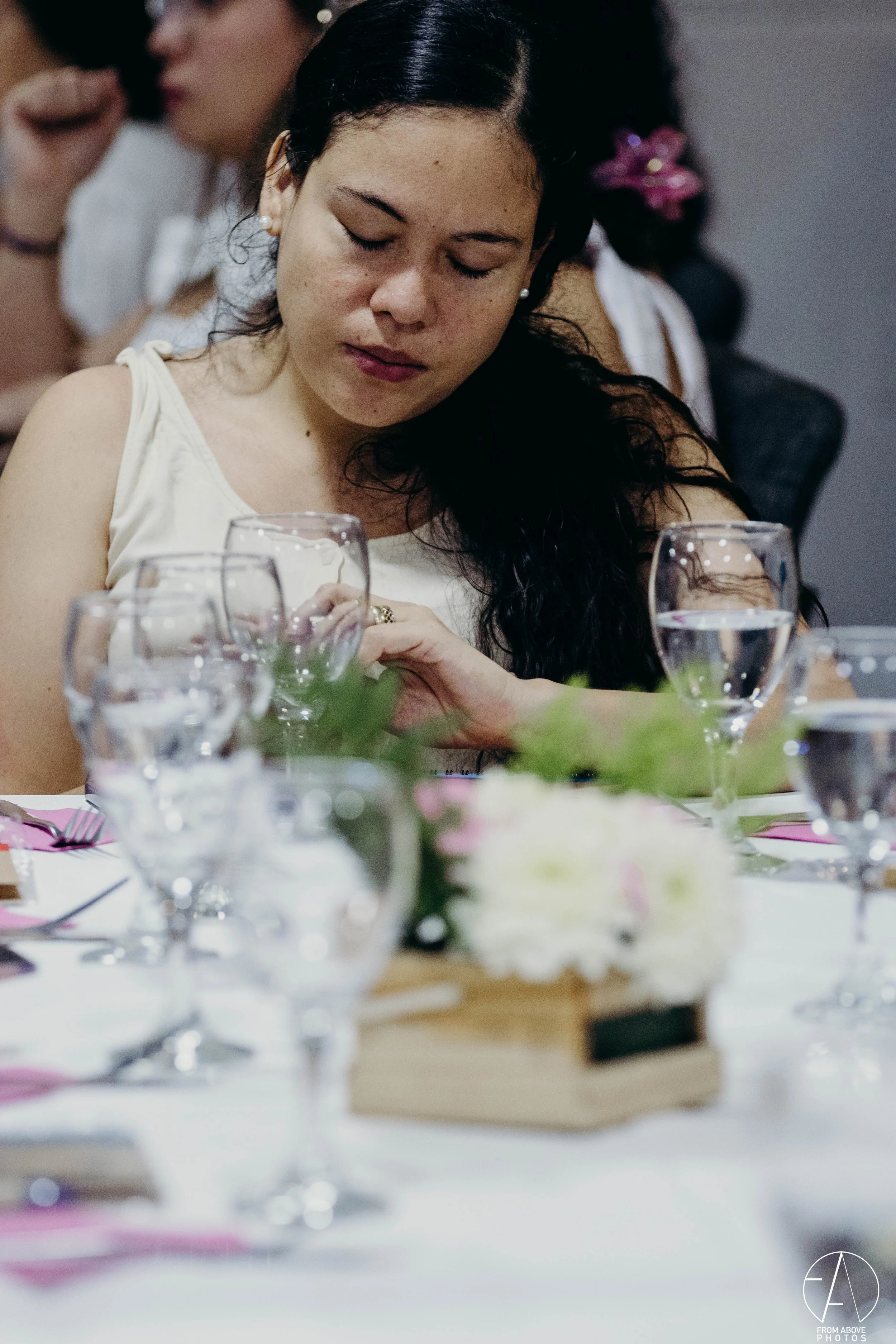 Mujer cerrando los ojos en una mesa con copas de agua y elementos decorativos, parece estar en una comida o evento formal.