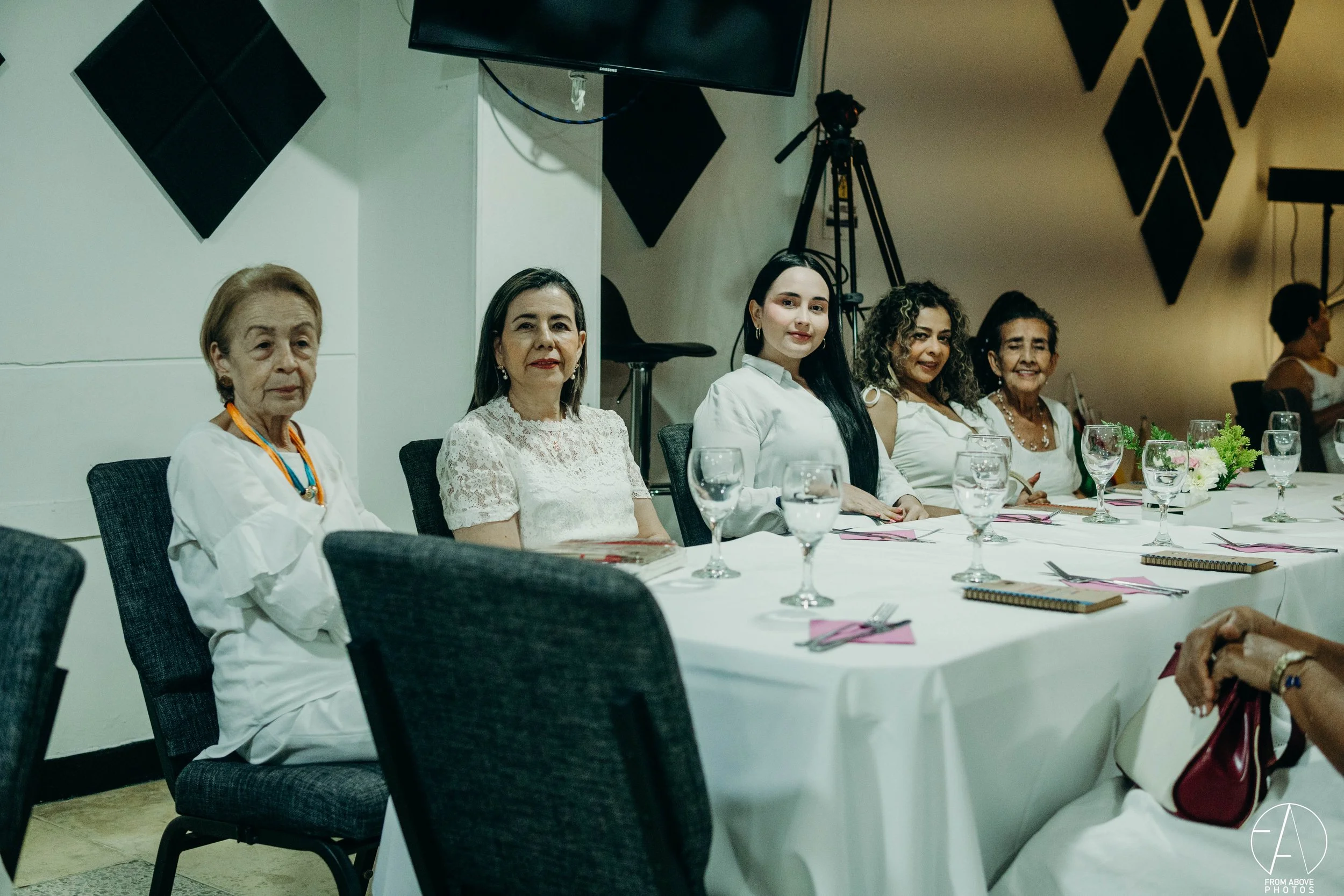 Grupo de seis mujeres sentadas en una mesa durante un evento, con copas de agua y papelitos rosados, en un entorno interior con paredes blancas y equipo de grabación en el fondo.