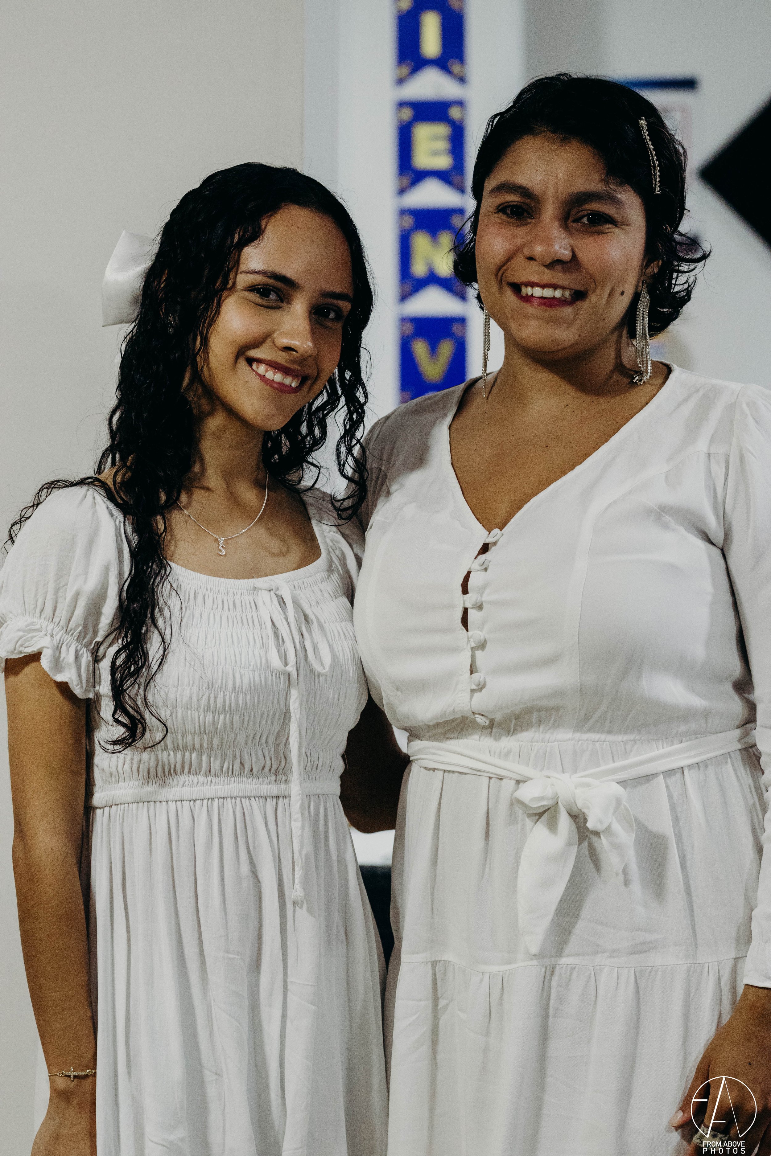 Dos mujeres vestidas con ropa blanca, sonriendo y posando juntas en un ambiente interior, con letras en la pared de fondo que parecen formar palabras en inglés.