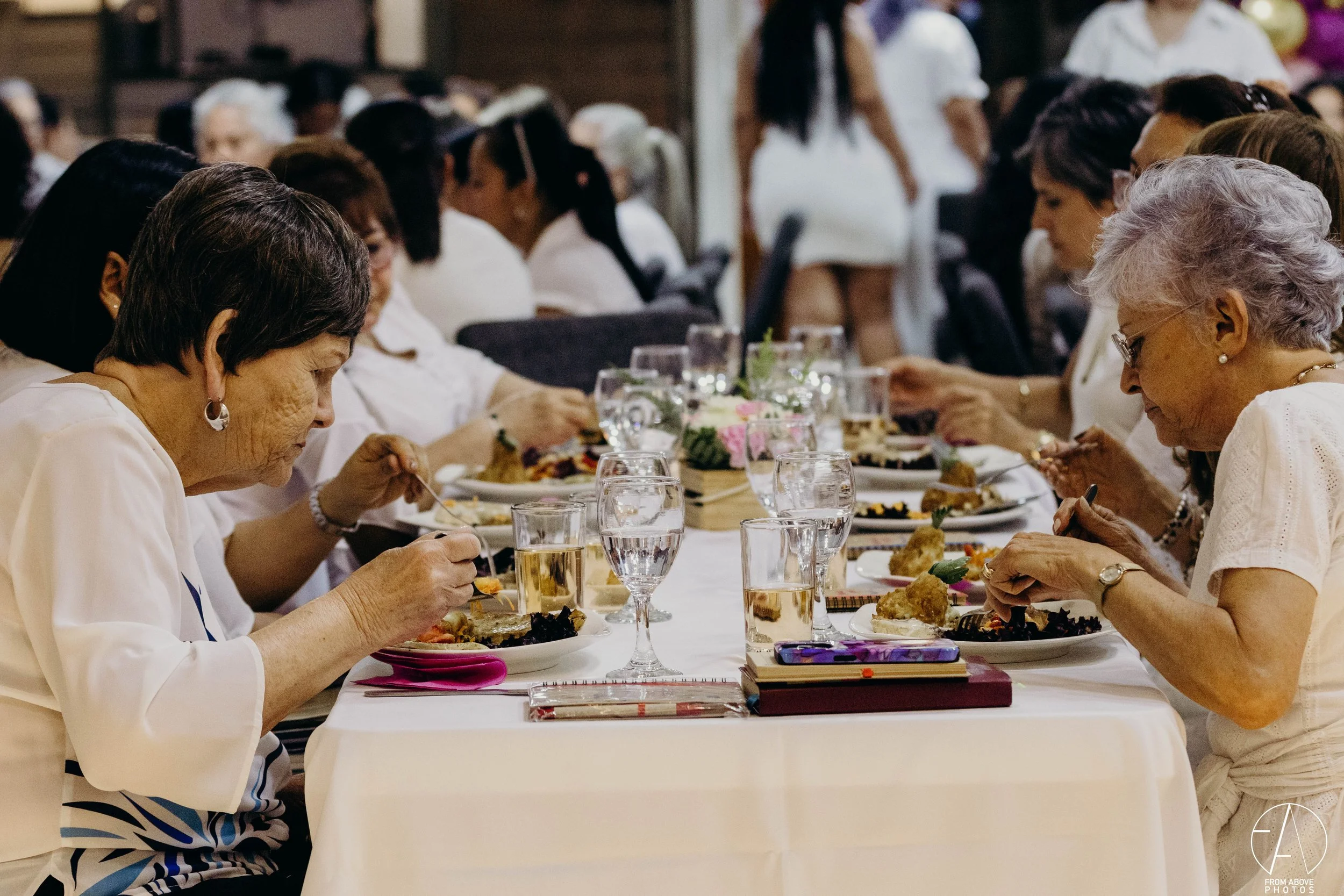 Un grupo de personas mayores, en su mayoría mujeres, sentadas en una mesa larga disfrutando de una comida en un evento social o familiar.