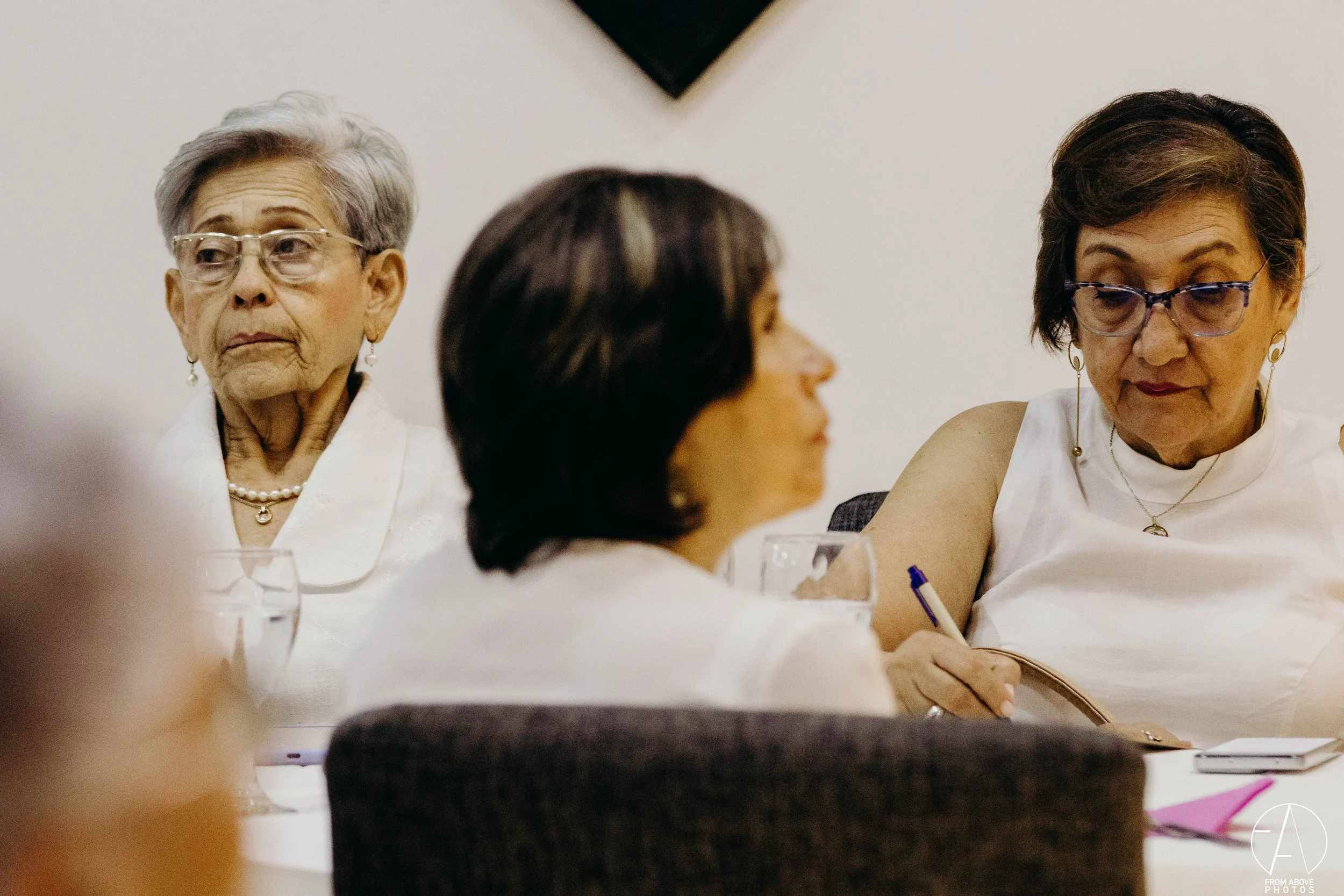 Tres mujeres adultas en una reunión, una de ellas cabizbaja y tomando notas.