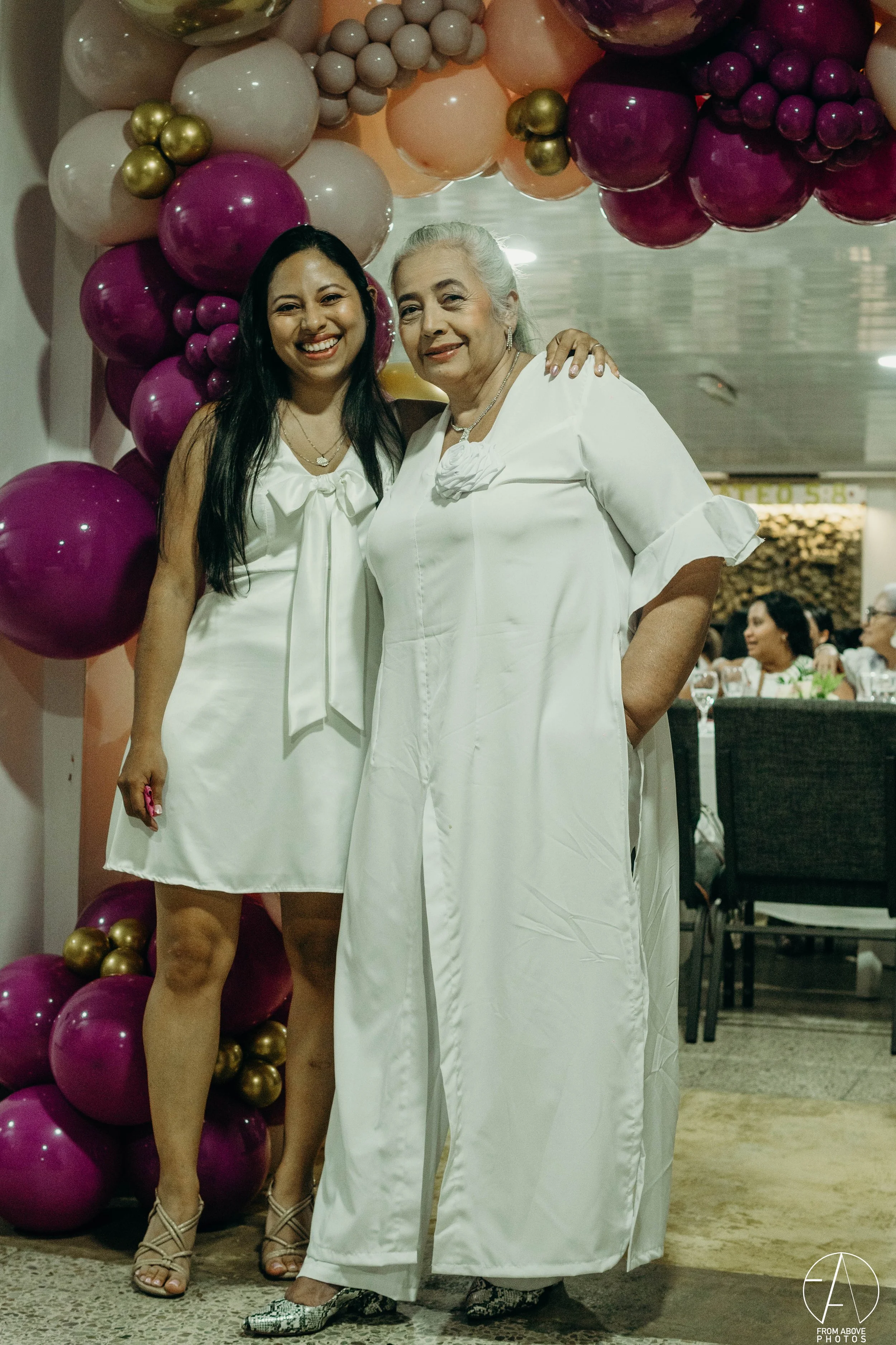 Dos mujeres vestidas de blanco, una joven y una mayor, sonriendo y abrazadas en un evento con decoraciones de globos en tonos rosados, dorados y beiges.