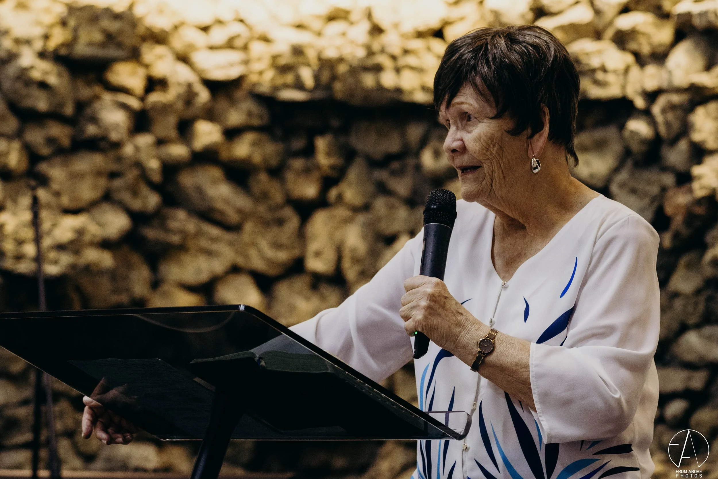 Anciana hablando en público con micrófono, sosteniendo un recetario, fondo de pared de piedra.