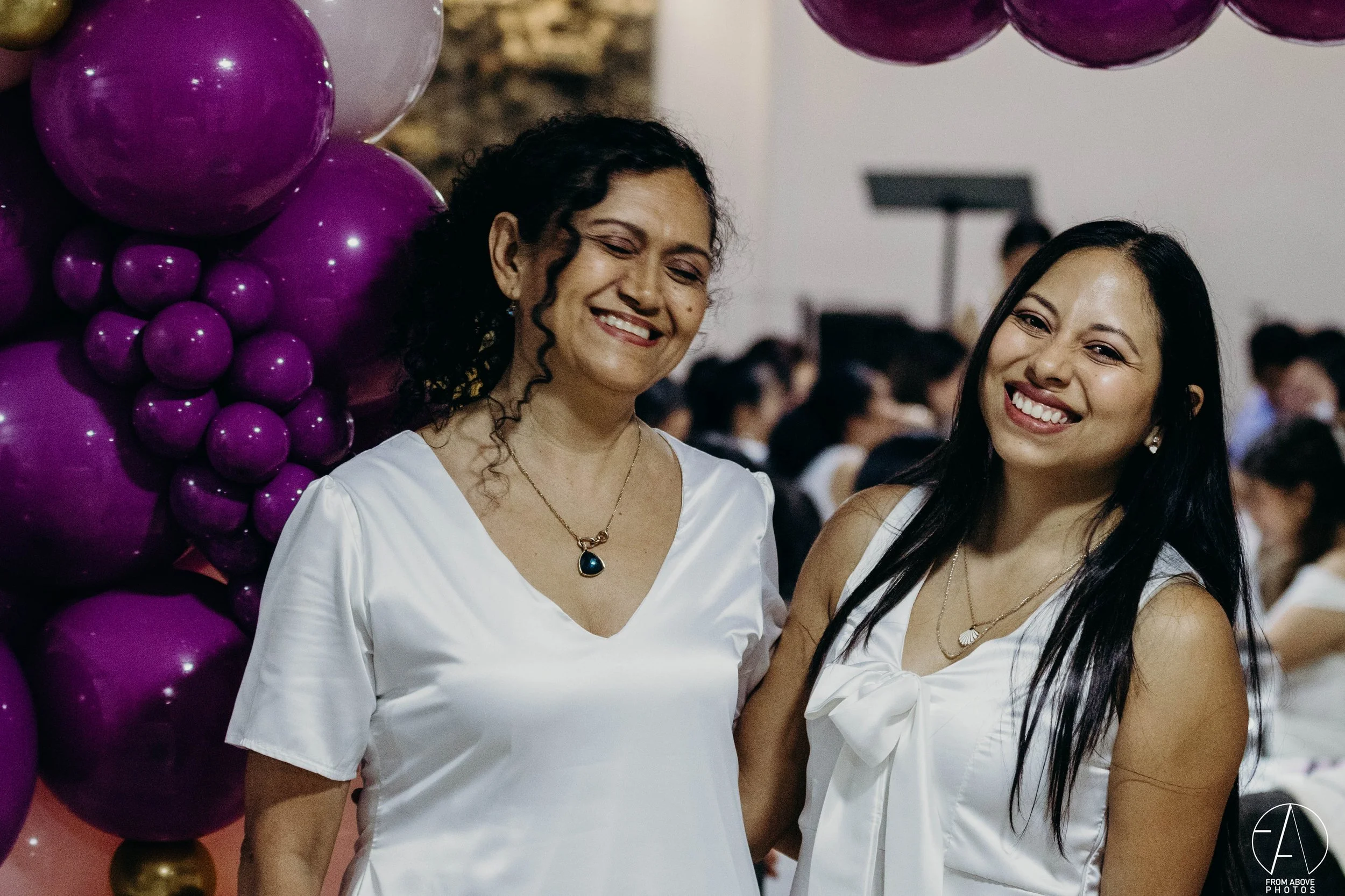 Dos mujeres sonriendo en una celebración, con decoración de globos morados y rosados en el fondo.