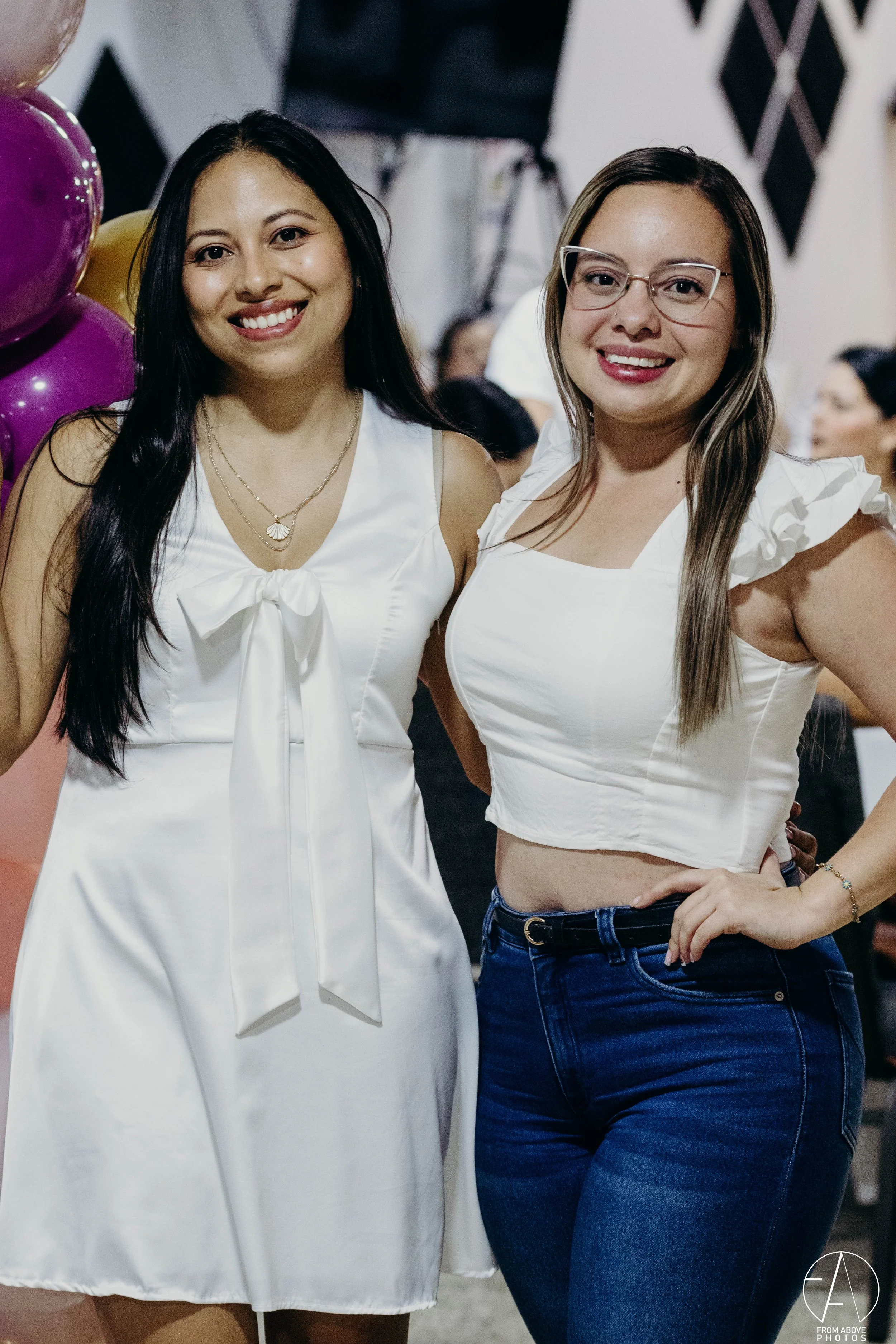 Dos mujeres sonriendo en una fiesta, una lleva un vestido blanco y la otra blusa blanca y jeans azules.