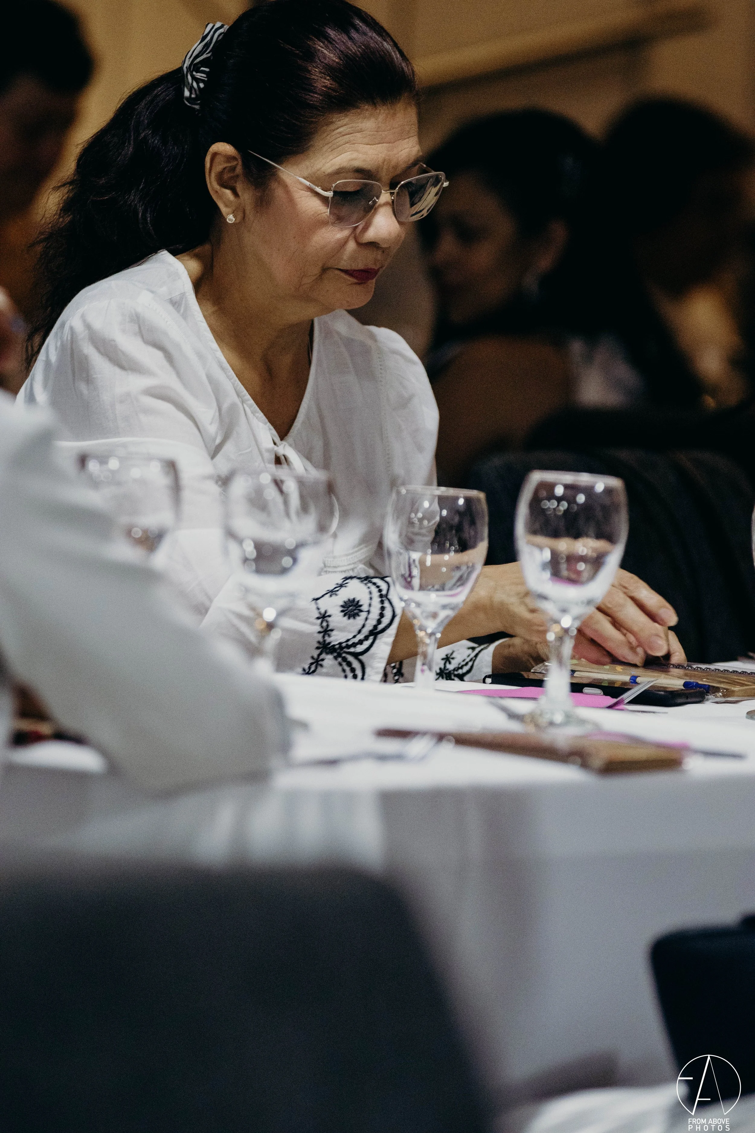 Mujer mayor con gafas y blusa blanca con bordados negros, sentada en una mesa en un evento formal. Frente a ella hay varias copas con agua y objetos de papelería.