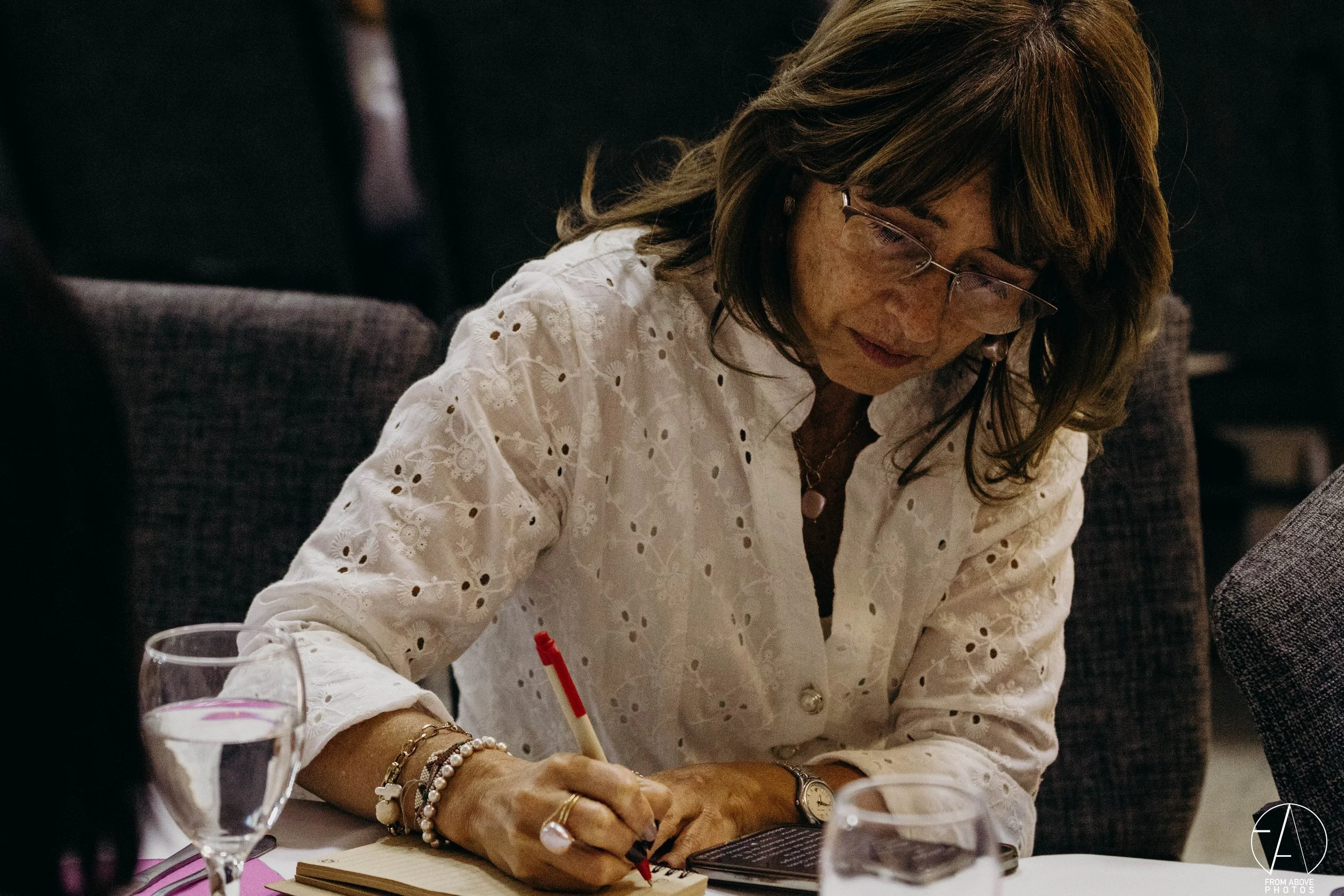 Mujer con gafas y cabello castaño, usando blusa blanca con bordado, escribe en un cuaderno en un restaurante o cafetería.