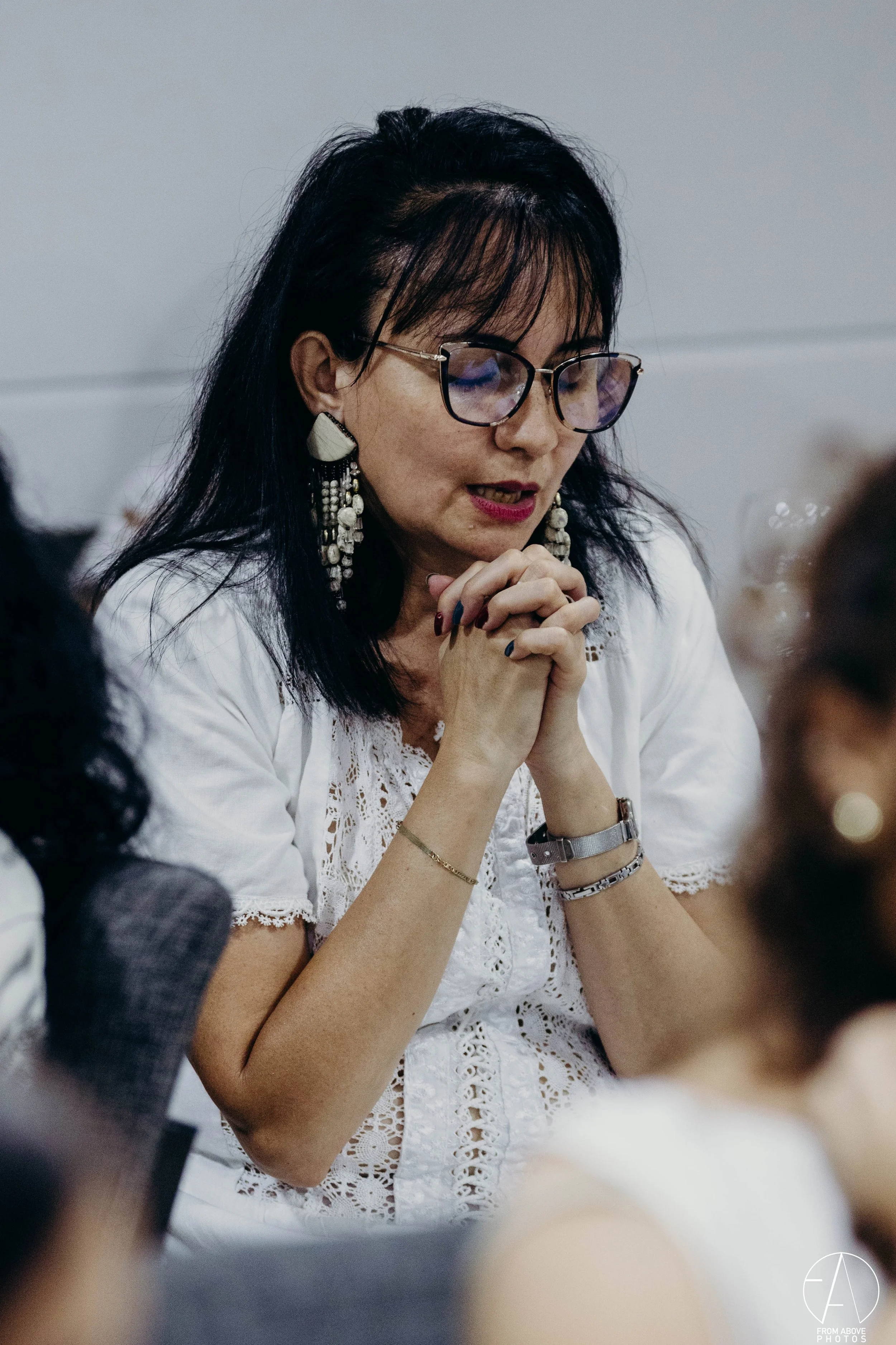 Mujer con gafas grandes y aretes largos, con las manos juntas en oración, vestida con blusa blanca con encaje.