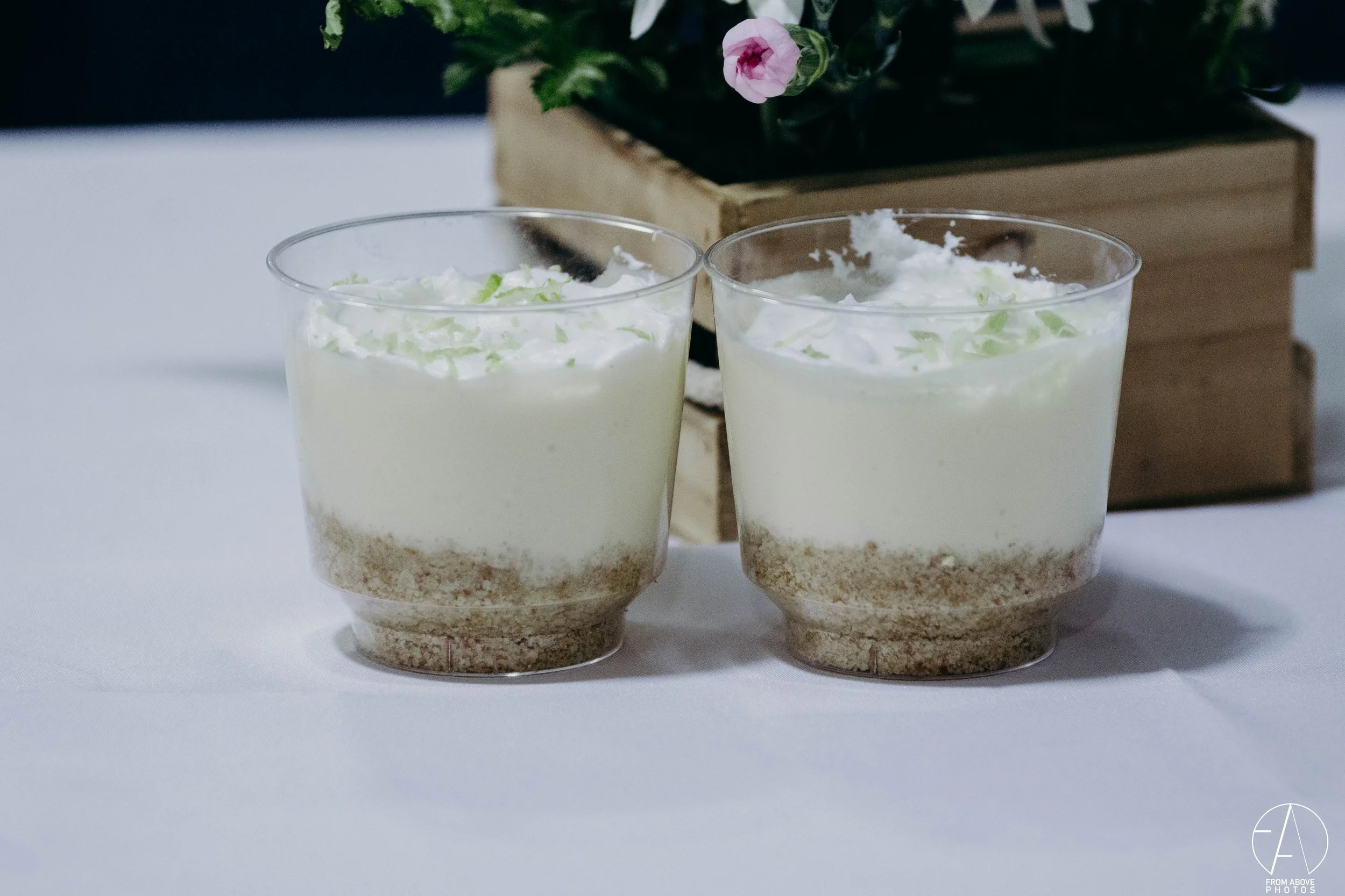Dos vasos con postre de gelatina y crema con virutas verdes decorativas, sobre una mesa blanca con fondo de cajas de madera y planta con flores rosas.