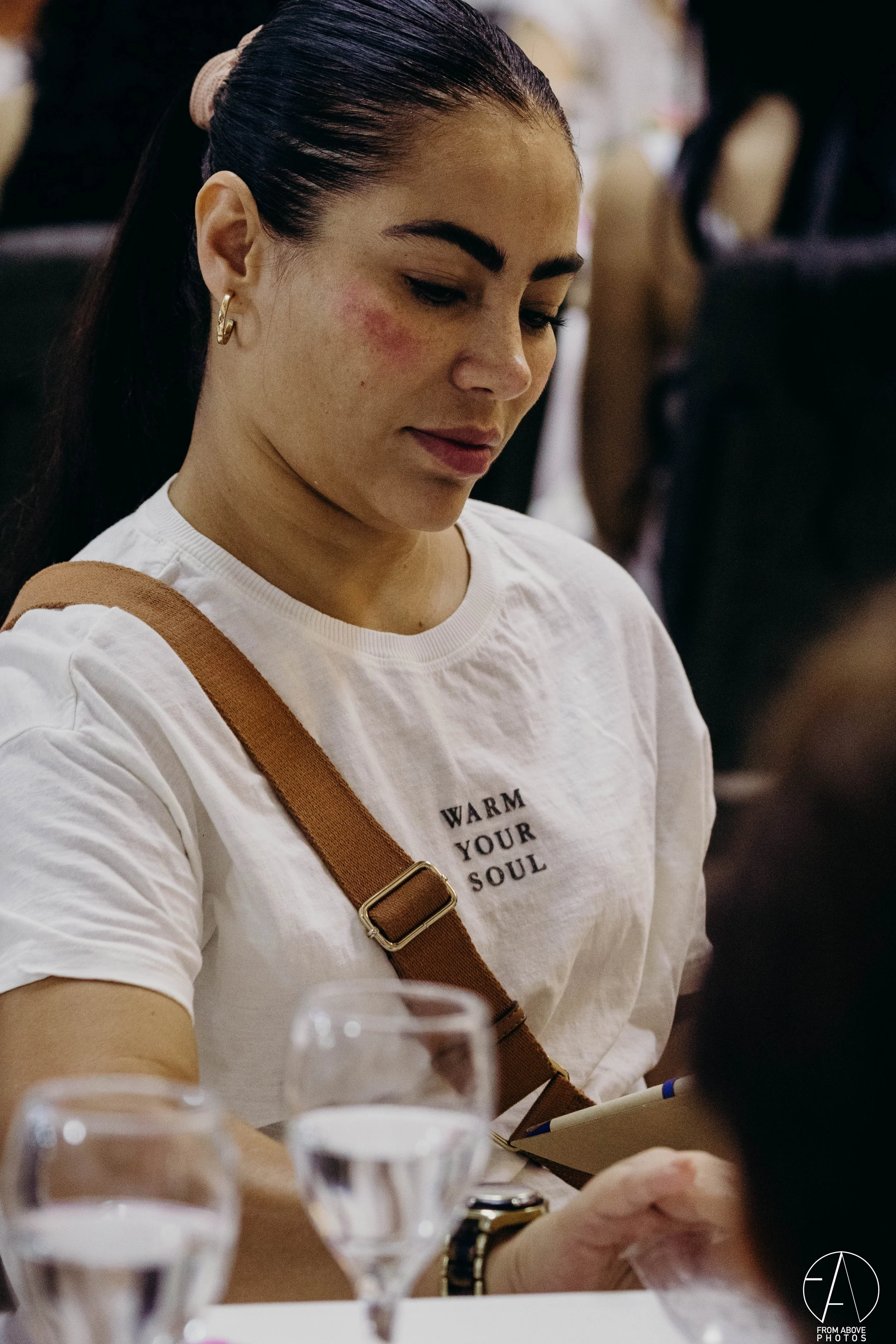 Mujer en una reunión, lleva una camiseta blanca que dice 'Warm Your Soul', con una mochila marrón y un reloj.