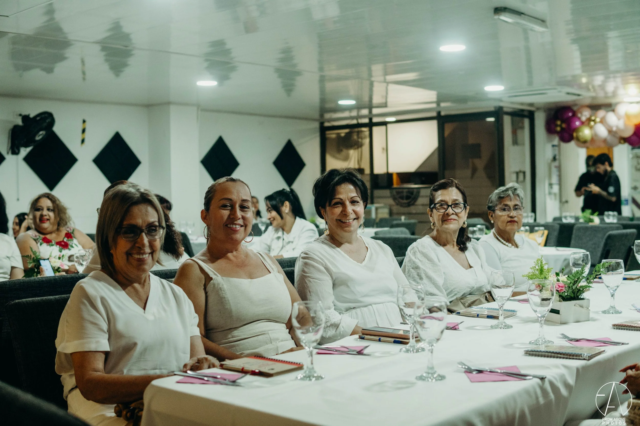 Grupo de mujeres sentadas en una mesa decorada con flores, vasos y cuadernos en un evento social en un salón con decoraciones en el fondo.