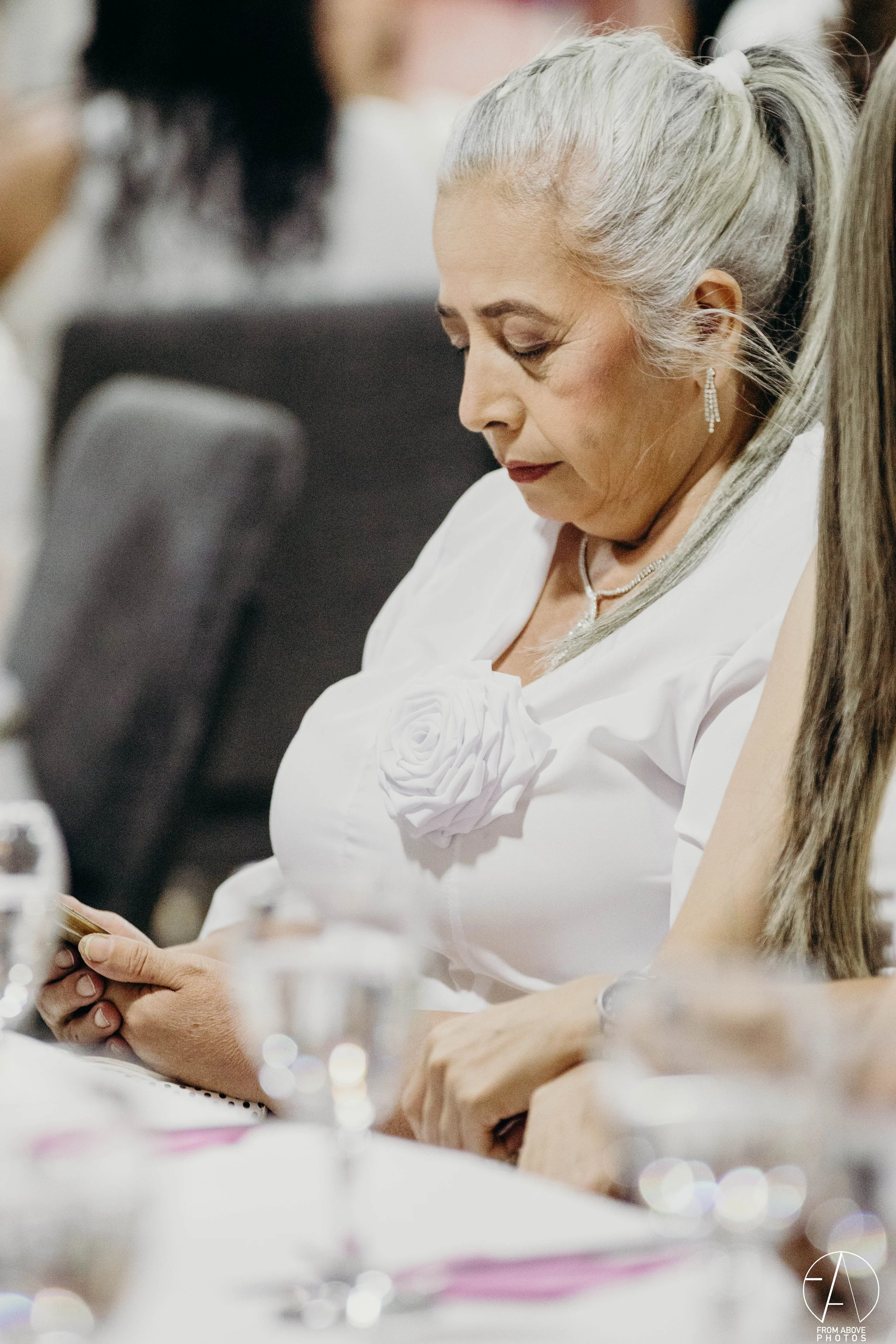 Mujer de cabello plateado atenta en una reunión o evento, usando ropa blanca con detalle de rosa en el pecho, y joyería, sentada en una mesa con bebidas y objetos en el fondo.