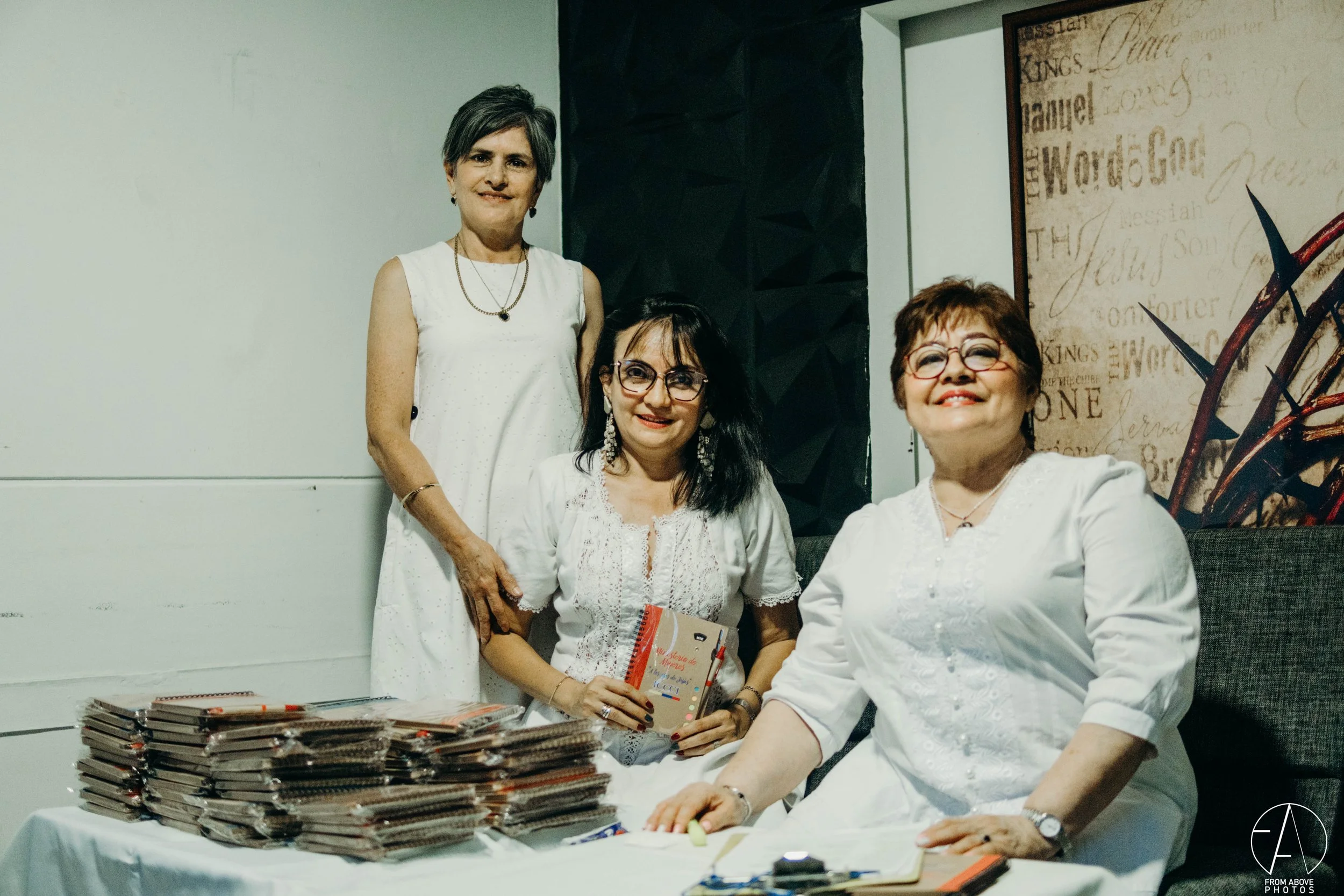Tres mujeres en una reunión, dos están sentadas y una de pie, con una pila de libros y papeles sobre la mesa