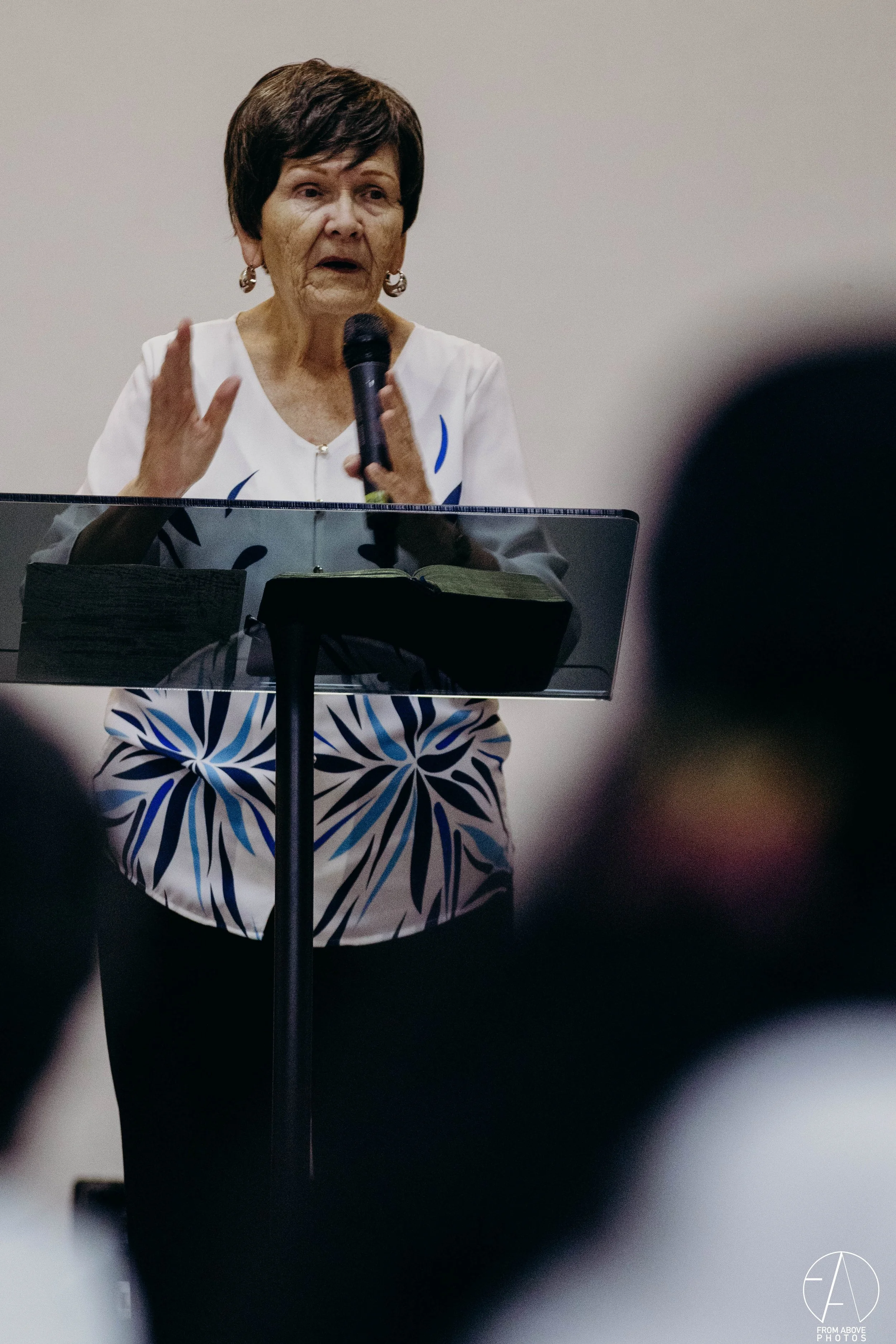 Mujer mayor hablando con micrófono en un podio, rodeada de personas en una conferencia o reunión.