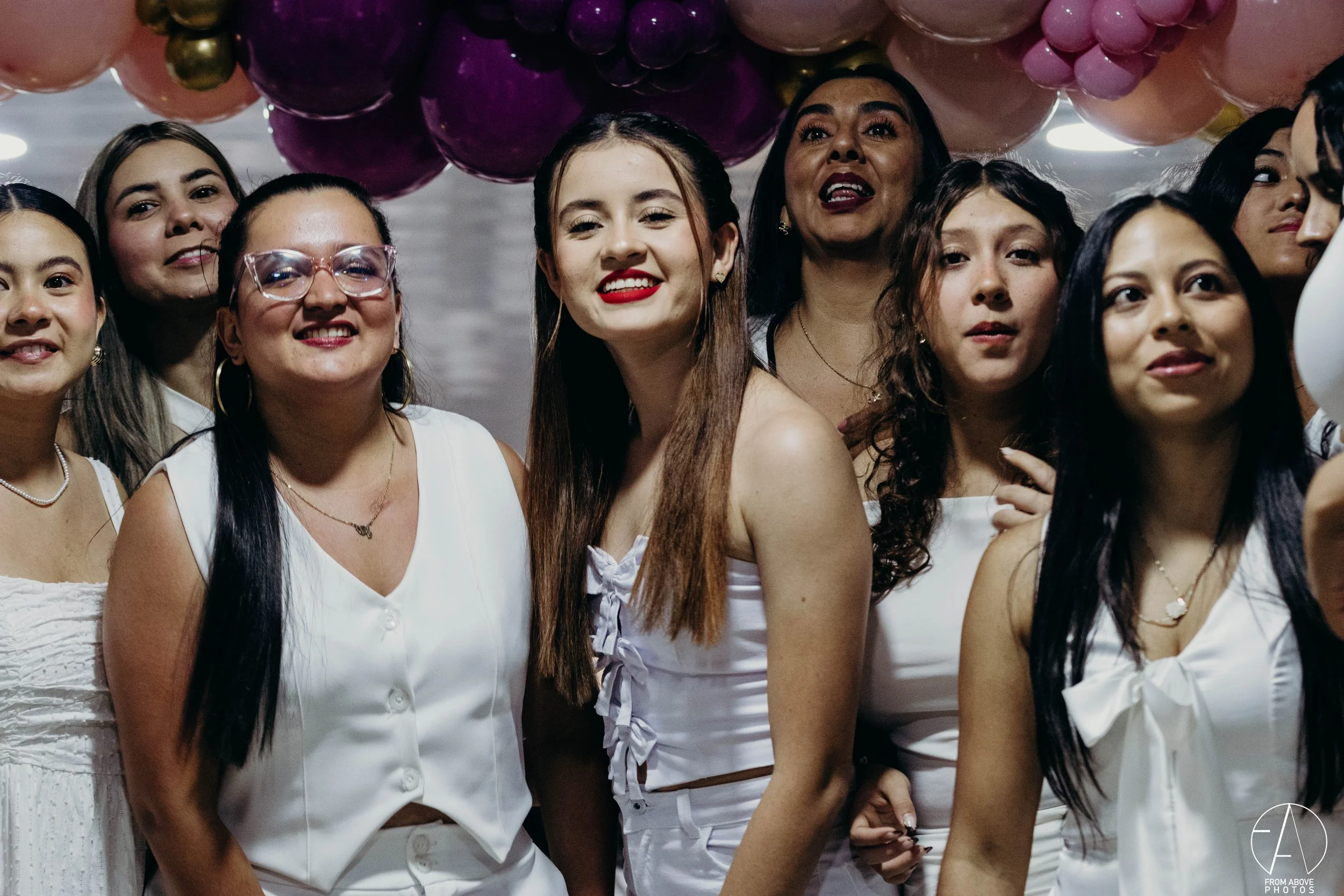 Grupo de mujeres en celebración con decoraciones de globos rosas y morados, sonriendo y vestidas con ropa blanca.