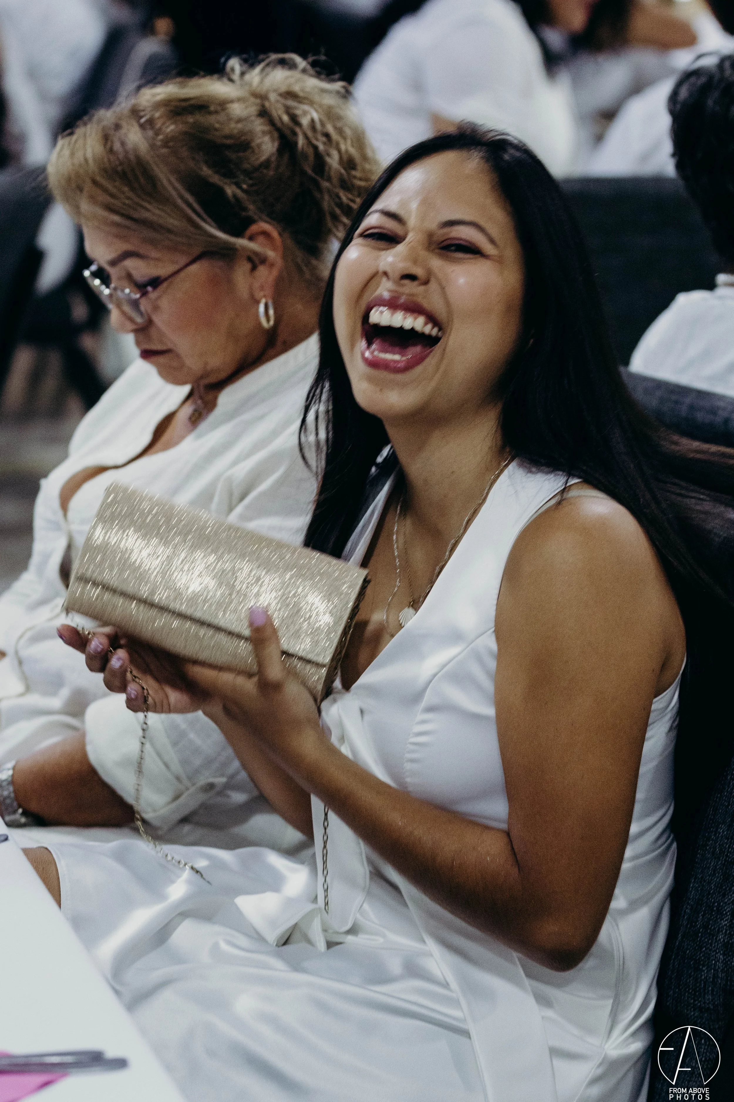 Mujer sonriente con vestido blanco, sosteniendo una cartera de mano dorada en un evento social, rodeada de otras personas.