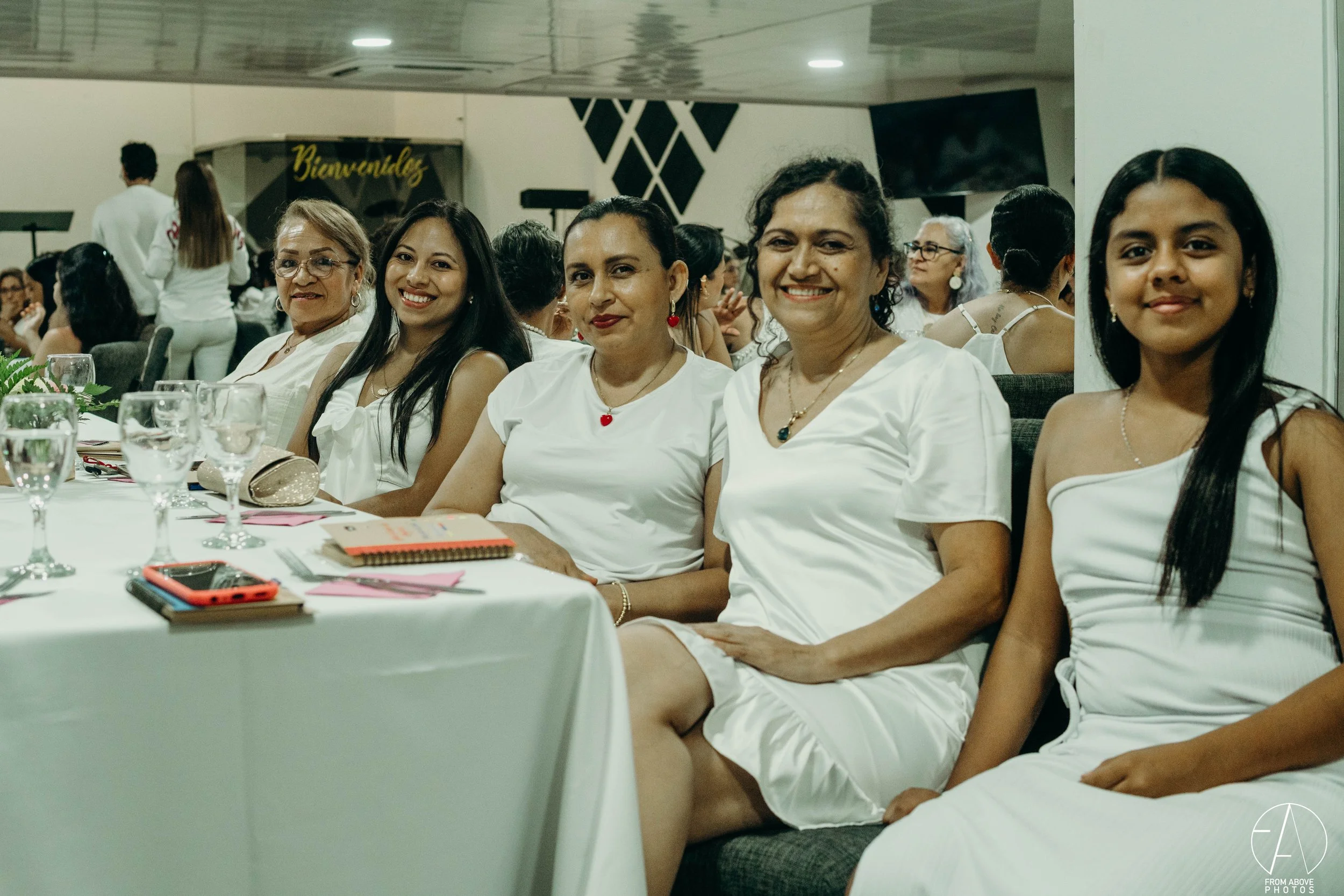 Grupo de seis mujeres y una niña sentadas en una mesa en un evento social, todas vestidas con prendas blancas y sonriendo.