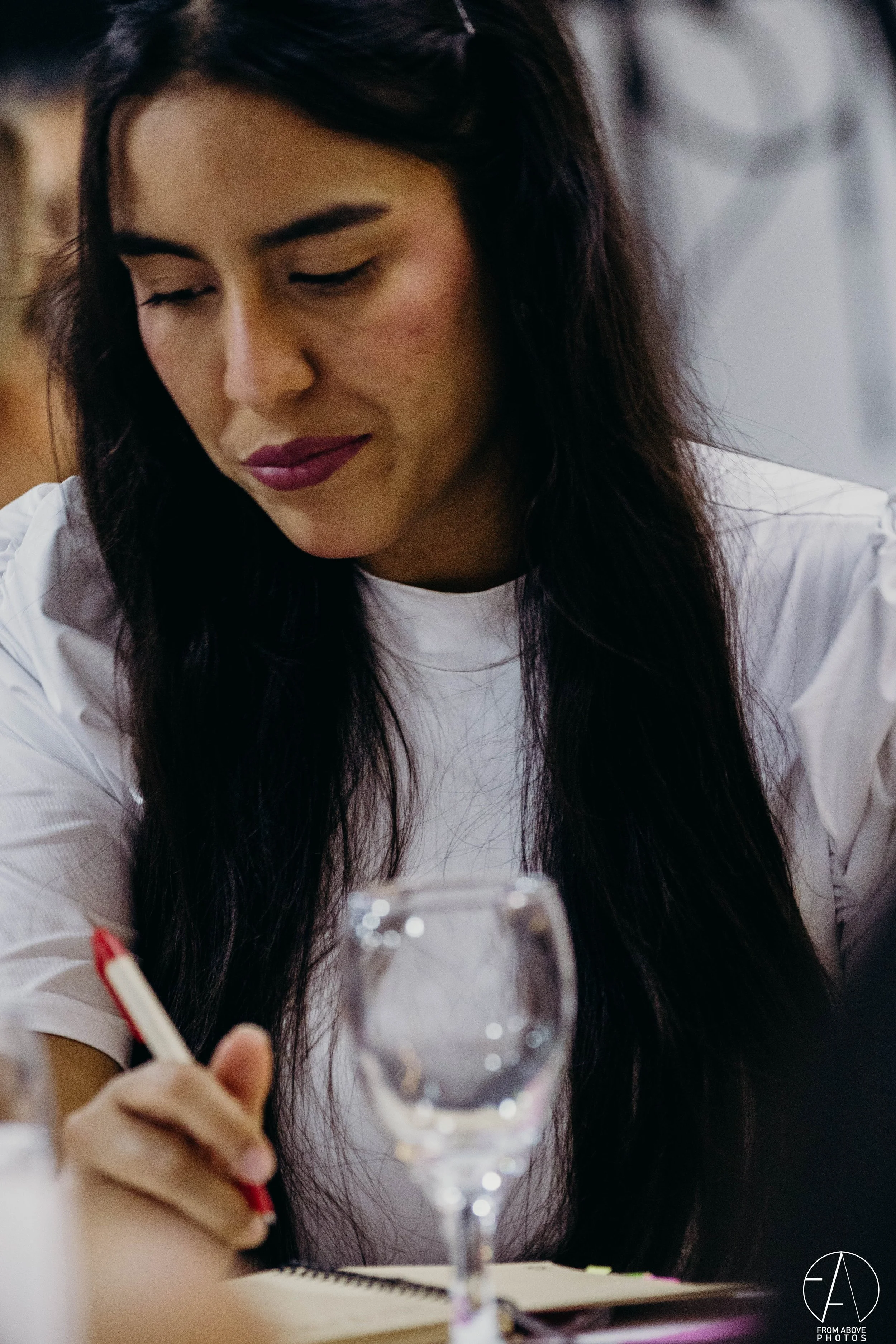 Mujer con cabello largo y oscuro, vestida con camisa blanca, escribiendo en un cuaderno con un bolígrafo rojo, en un entorno interior.