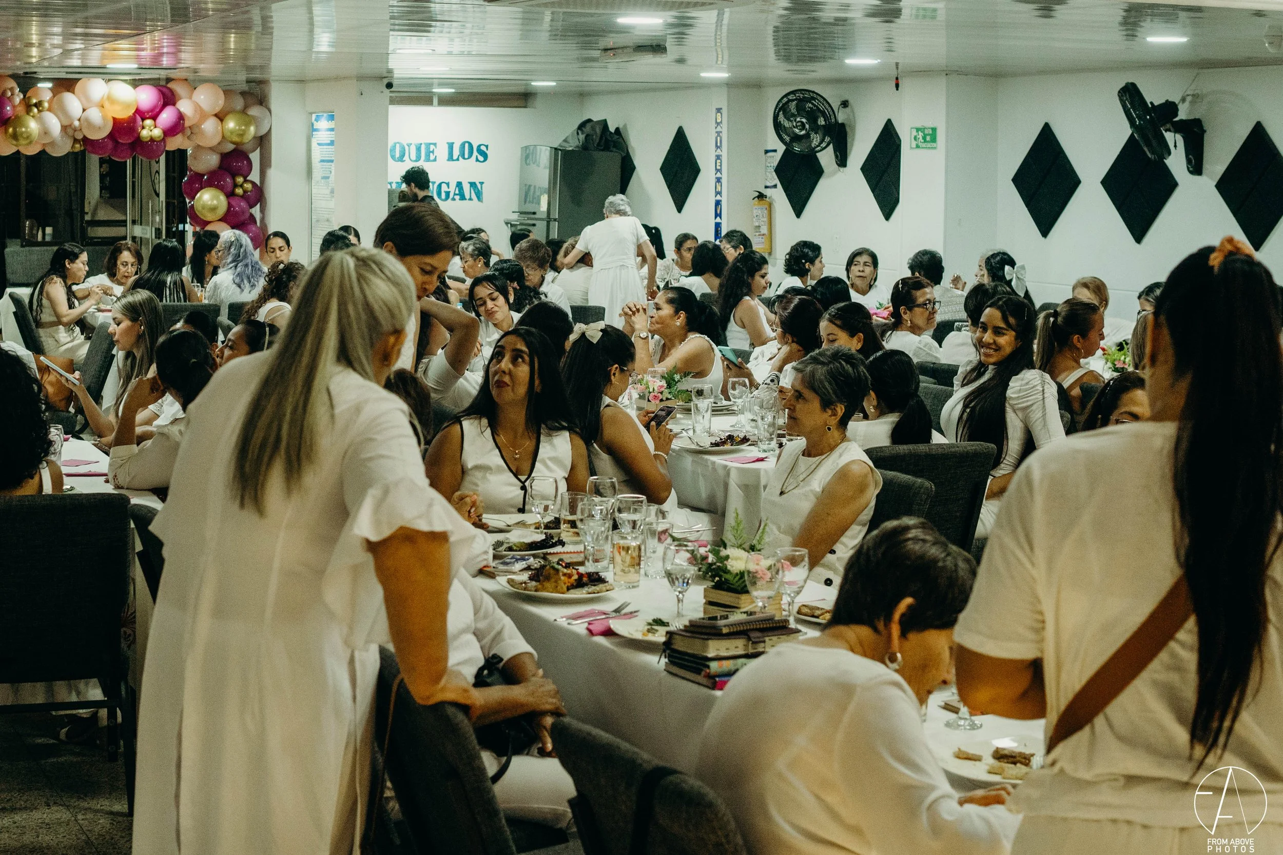 Muchísimas mujeres en una cena, la mayoría vestidas de blanco, en un lugar decorado con globos rosados y dorados, y mensajes en las paredes. Algunas personas están usando teléfonos móviles y hablando entre sí.