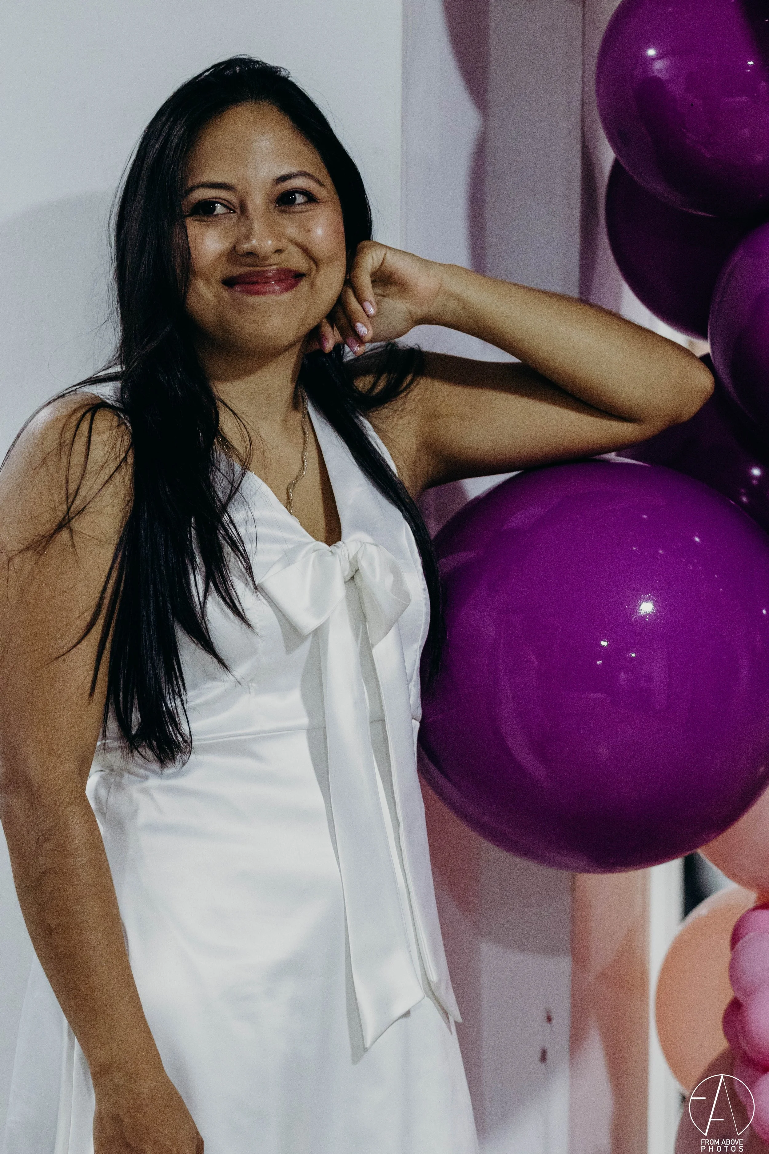 Mujer sonriendo con cabello largo y oscuro, vestida con vestido blanco con lazo en el cuello, rodeada de globos morados y rosados en un evento festivo.