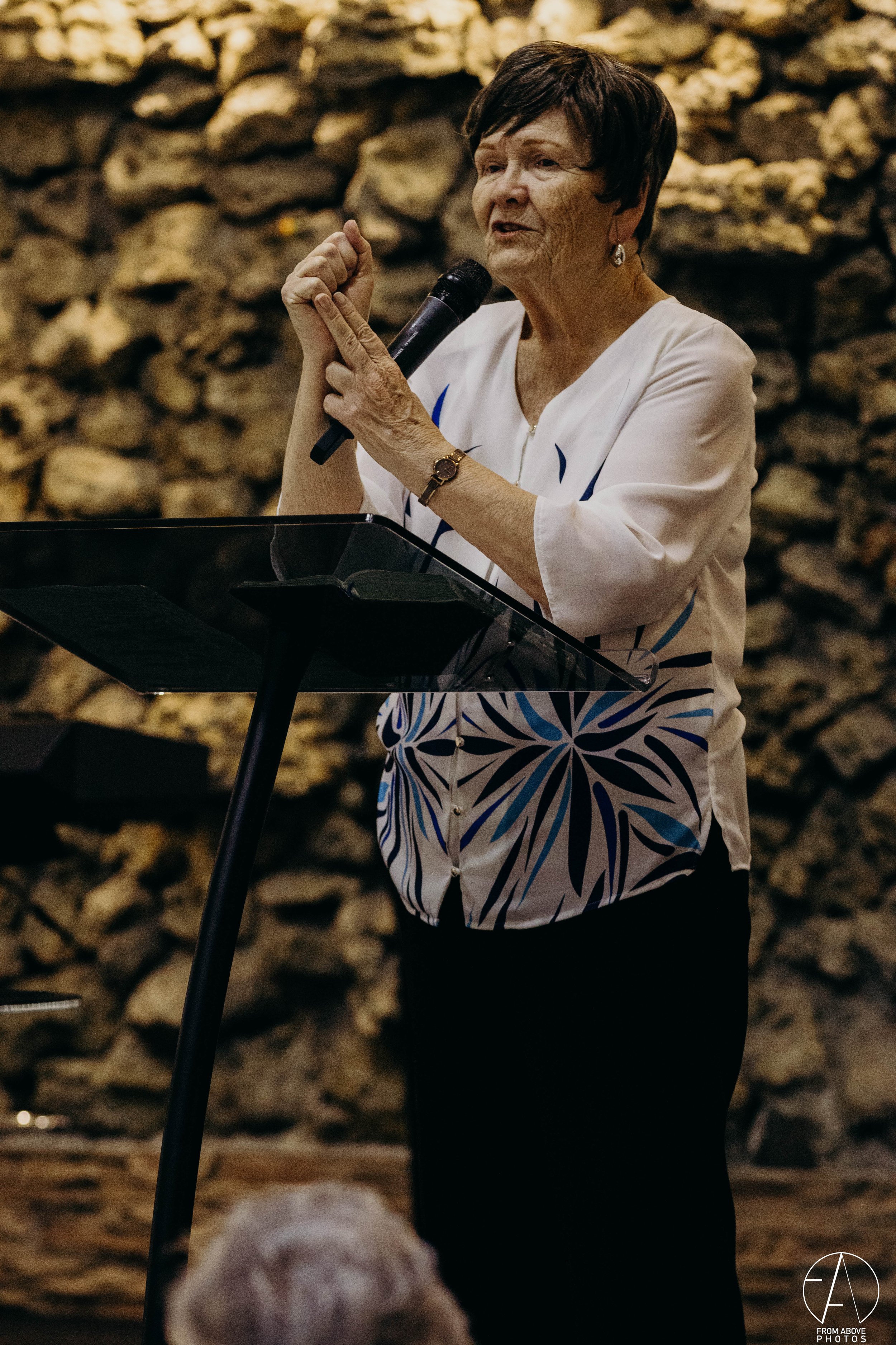 Una mujer anciana con cabello corto y oscuro habla en un micrófono, mientras está de pie frente a un atril en un entorno con muro de piedras en el fondo.