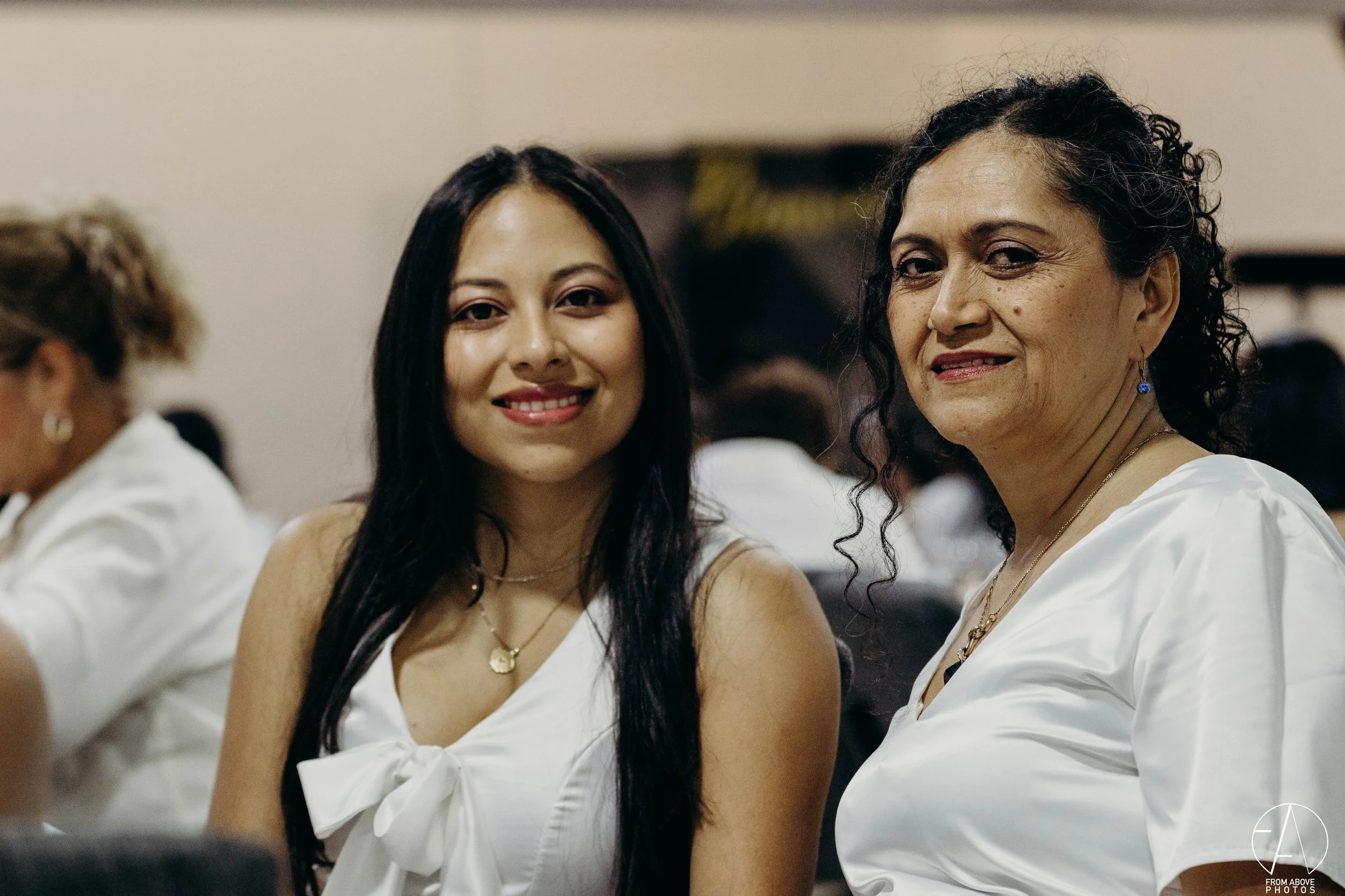 Dos mujeres sonriendo en un evento, una joven y una adulta mayor, vestidas con blusas blancas