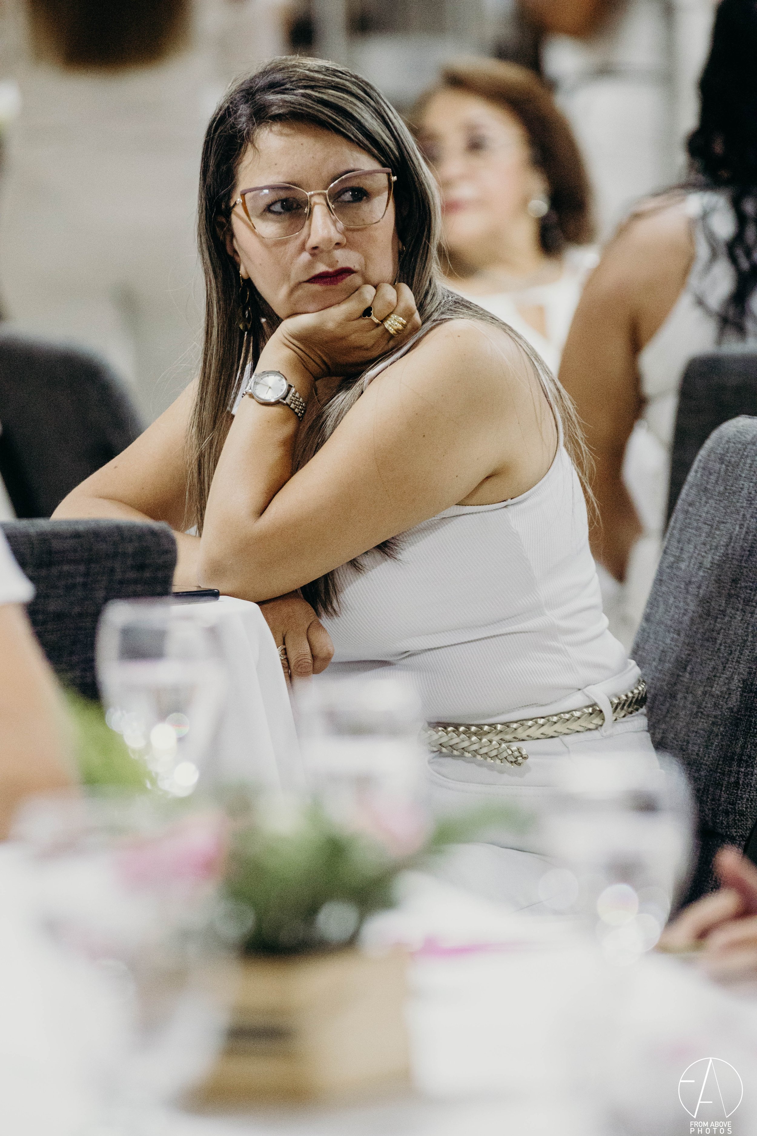 Mujer con gafas y ropa blanca, sentada en una mesa en un evento formal, con expresión pensativa y apoyo en la mano.