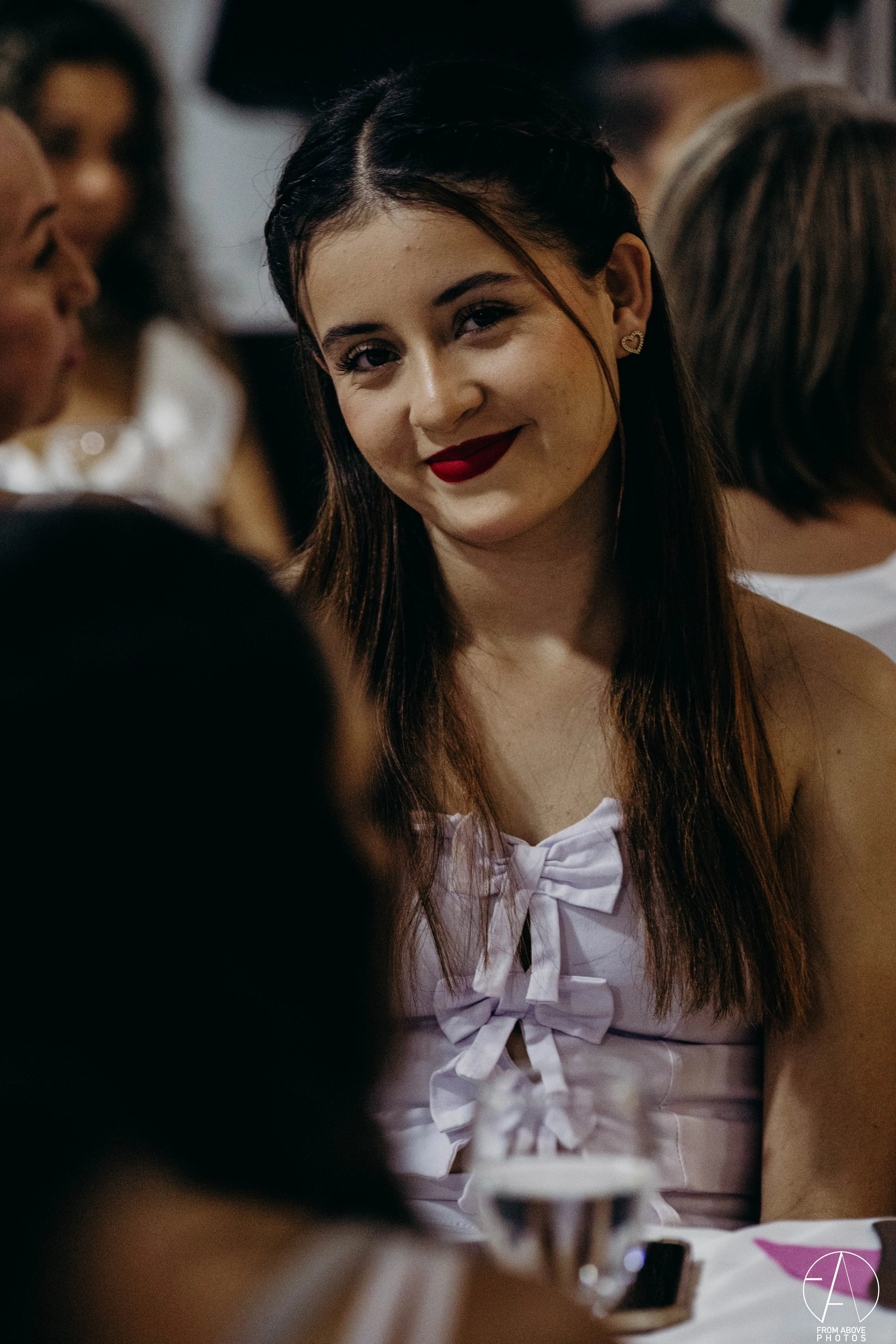Una joven con cabello largo y oscuro, con maquillaje llamativo, labial rojo y pendientes de corazón, vestida con blusa blanca con lazadas, en un evento social rodeada de otras personas.