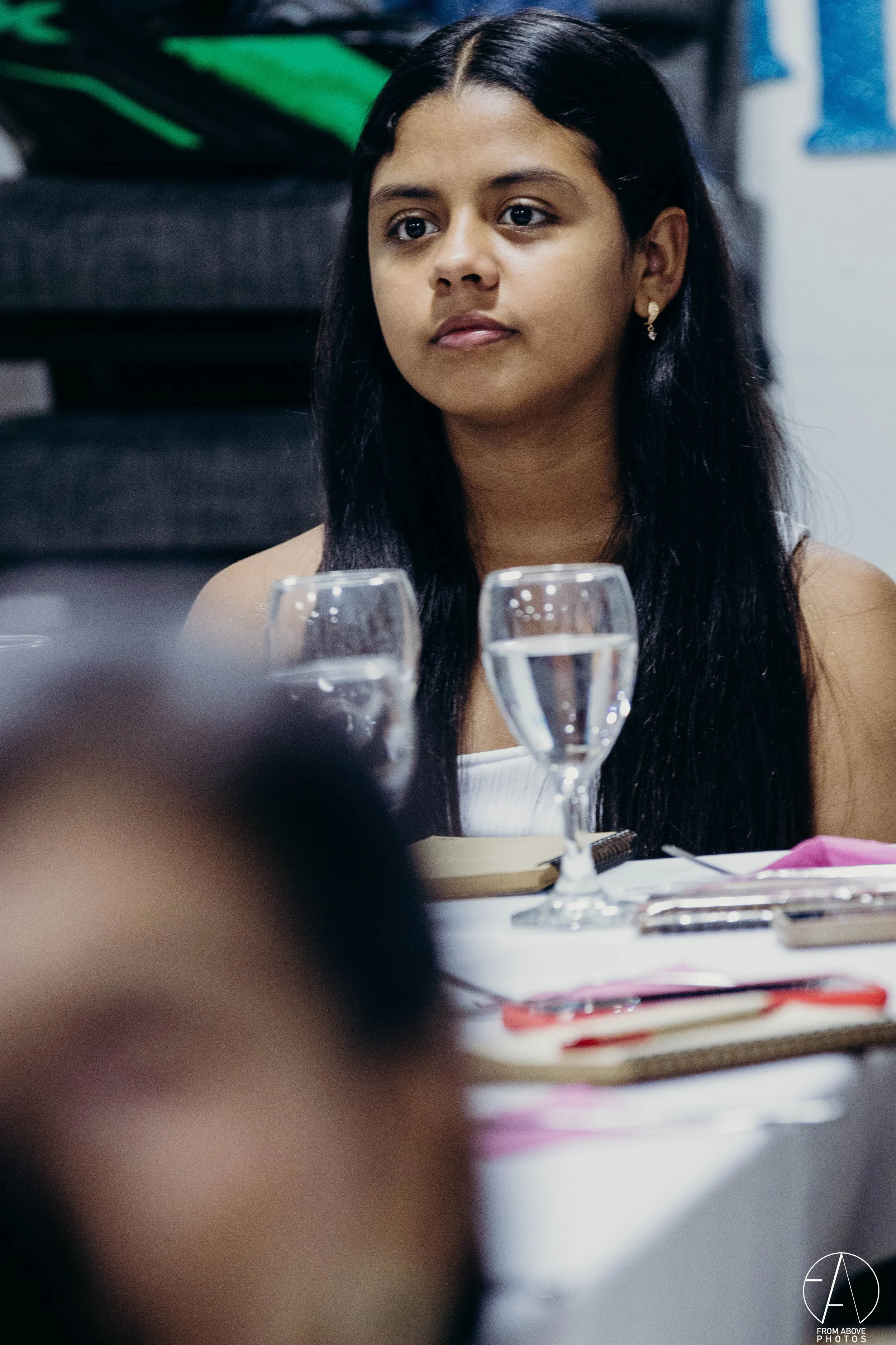 Joven mujer con cabello largo y oscuro, sentada en una mesa con dos copas de agua frente a ella, en un entorno interior.