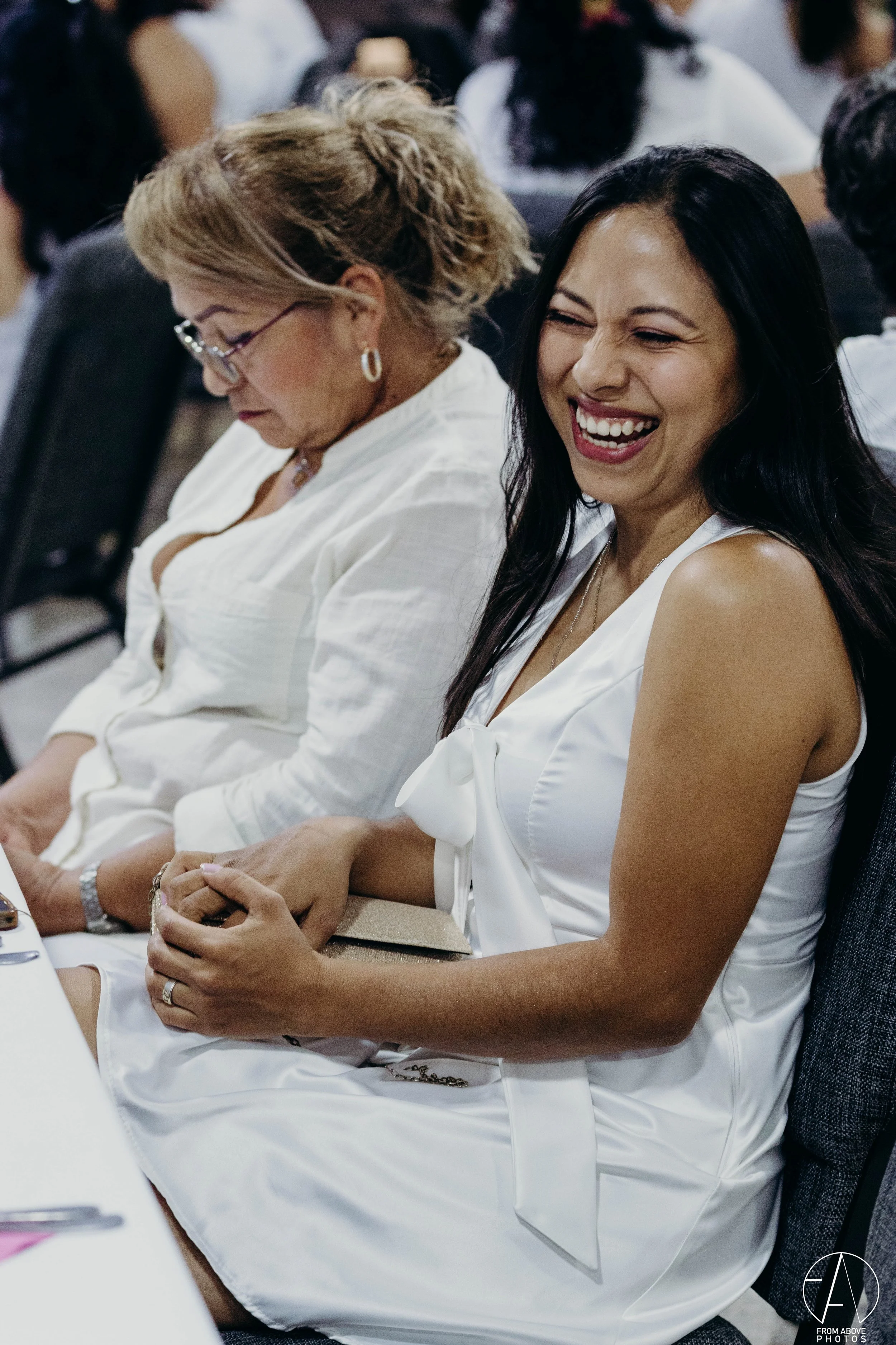 Mujer rubia con gafas y mujer de cabello negro sonriendo y sentadas en una fila en un evento, con muchas personas en el fondo.