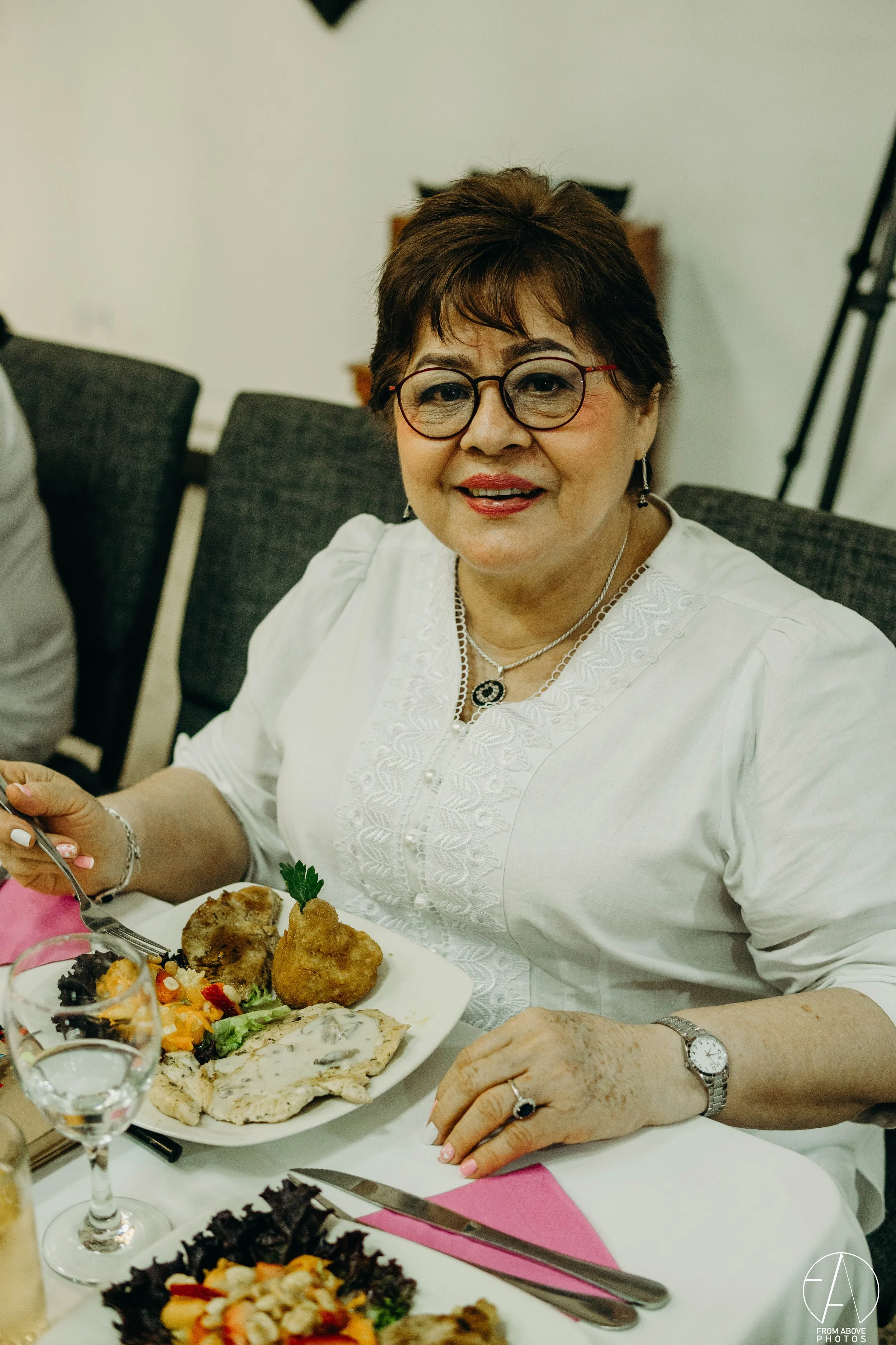 Mujer de cabello corto y gafas, sonriendo en una comida, con plato de comida que incluye pollo, ensalada y otros alimentos.