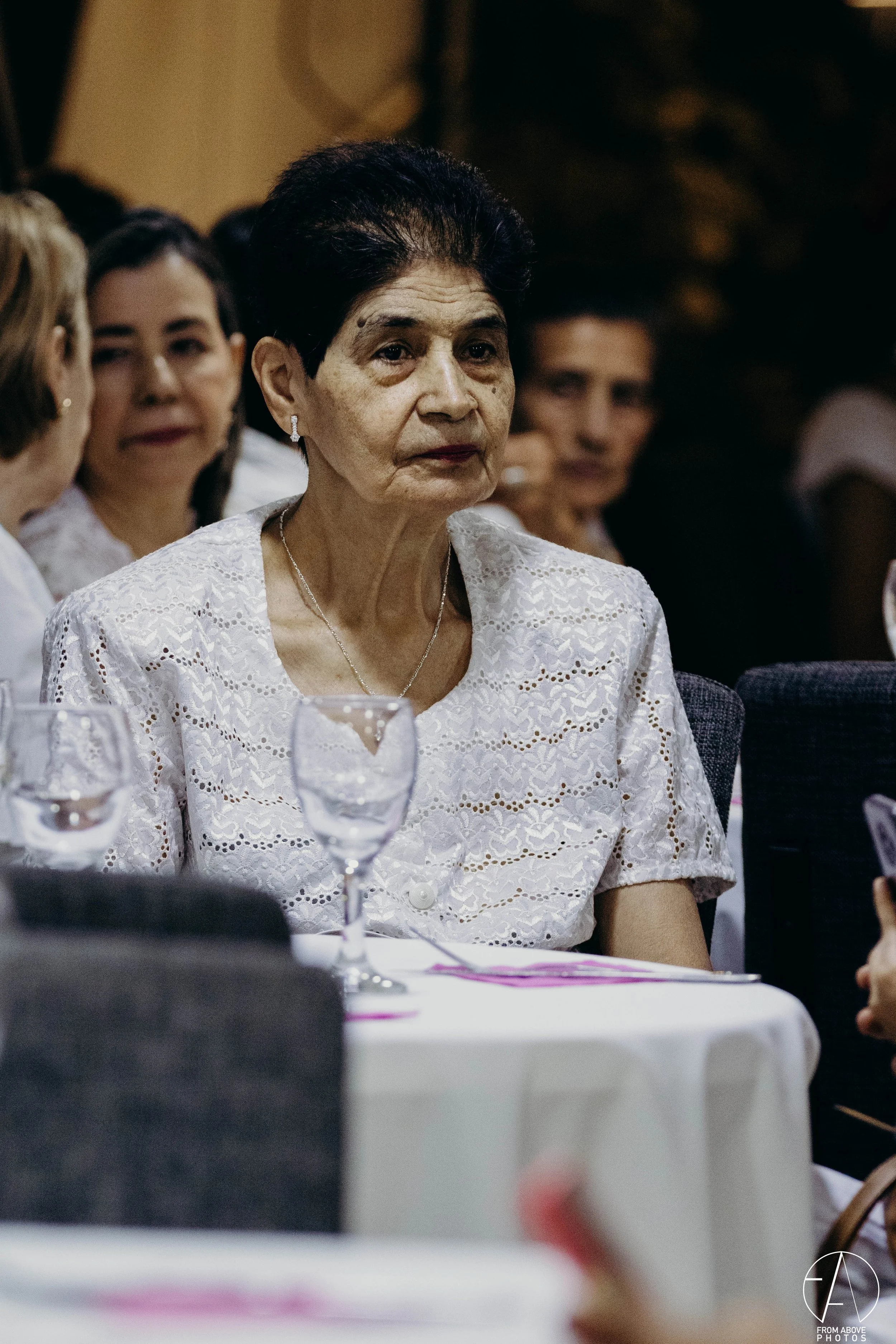 Una mujer de cabello oscuro y canoso vestida con blusa de encaje blanca en una reunión o evento formal, rodeada de otras personas.