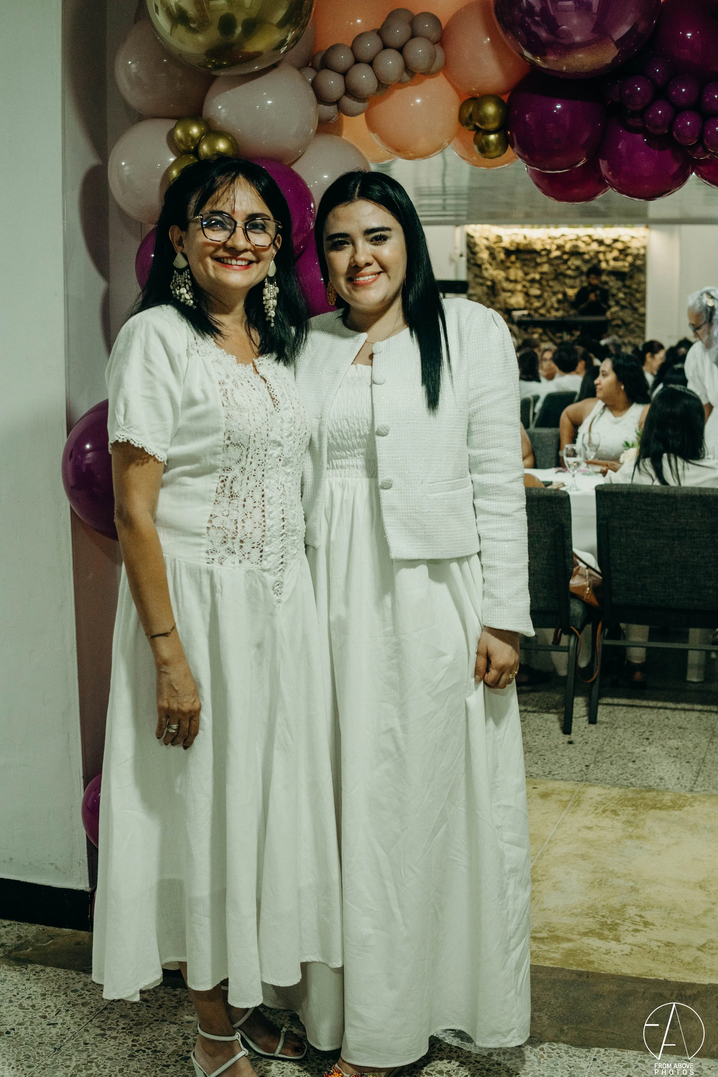 Dos mujeres en un evento social, vestidas con ropa blanca, con decoraciones de globos de colores en el fondo.