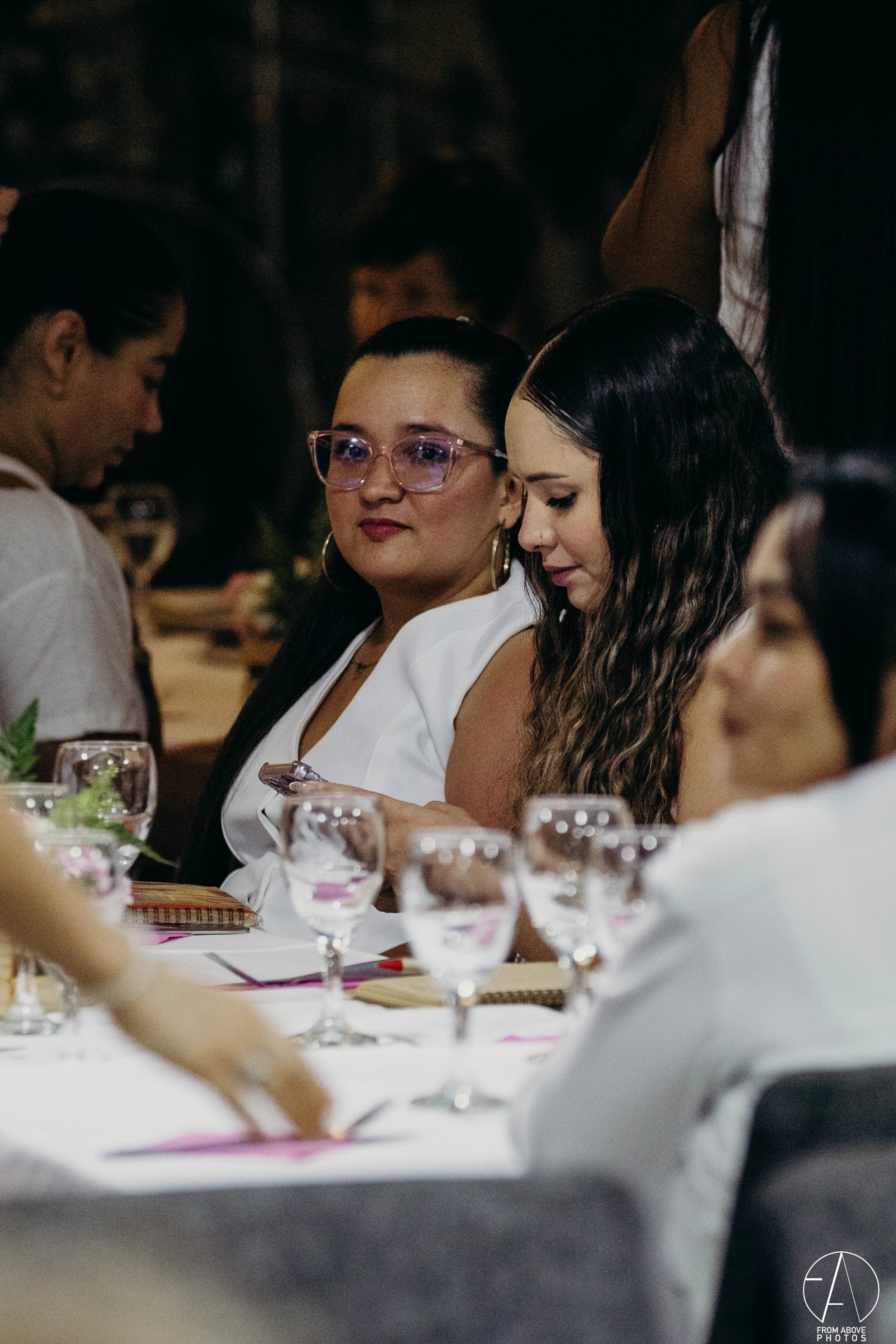 Mujeres en una reunión o evento social sentadas en una mesa con copas y decoraciones, una de ellas utilizando gafas de pasta y peinado liso, otra con cabello rizado y peinado oscuro.