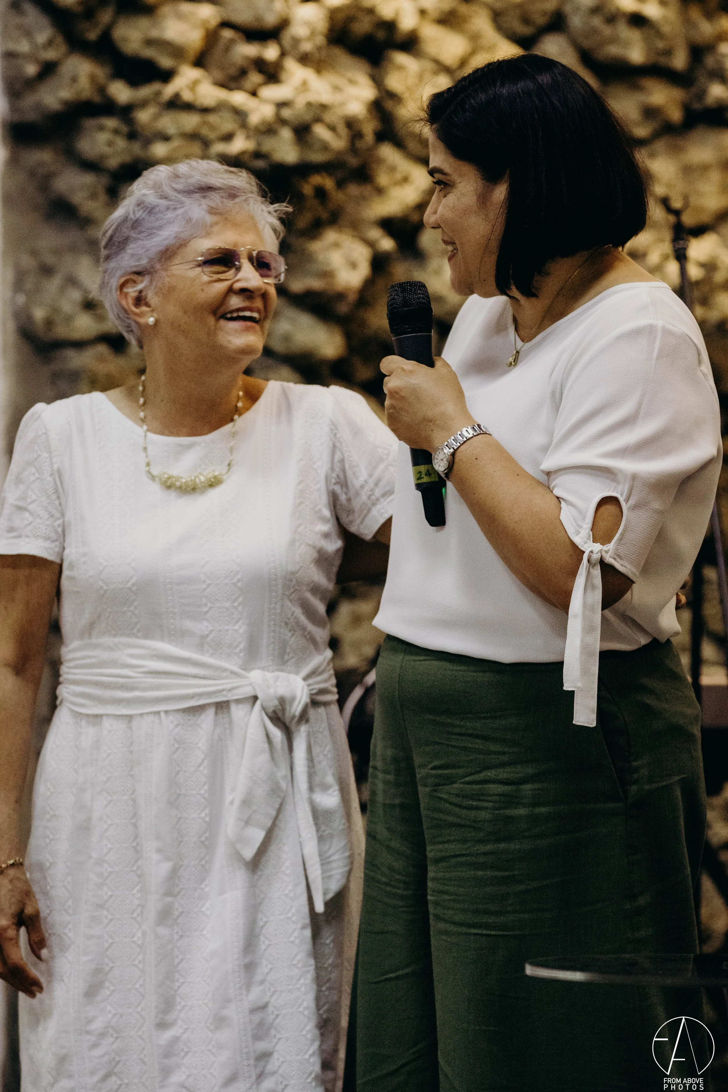Dos mujeres interactuando, una con un micrófono, celebrando en un ambiente con paredes de piedra.