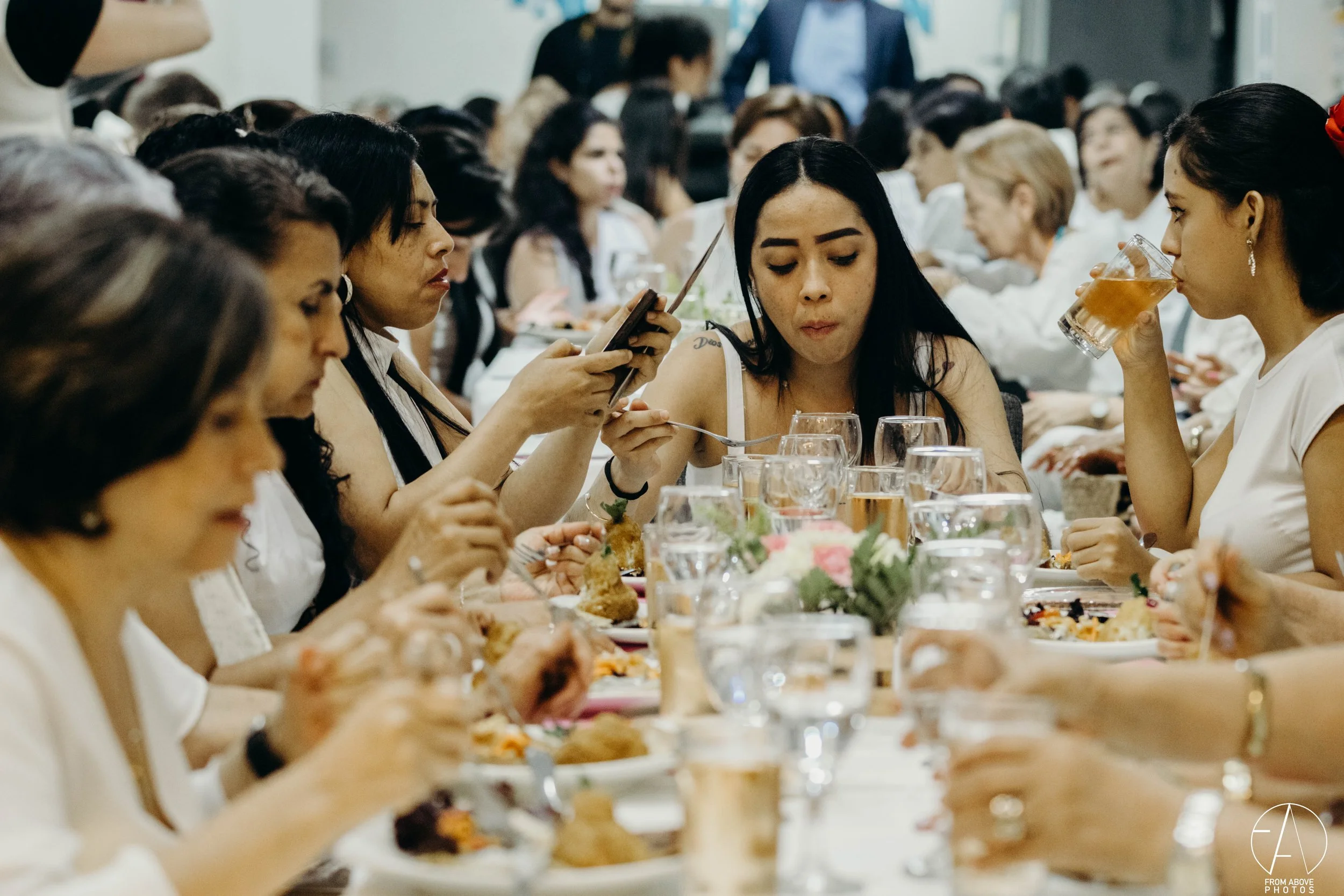 Varias mujeres sentadas en una mesa larga en una comida social, algunas con bebidas y comida en platos, en un entorno interior.