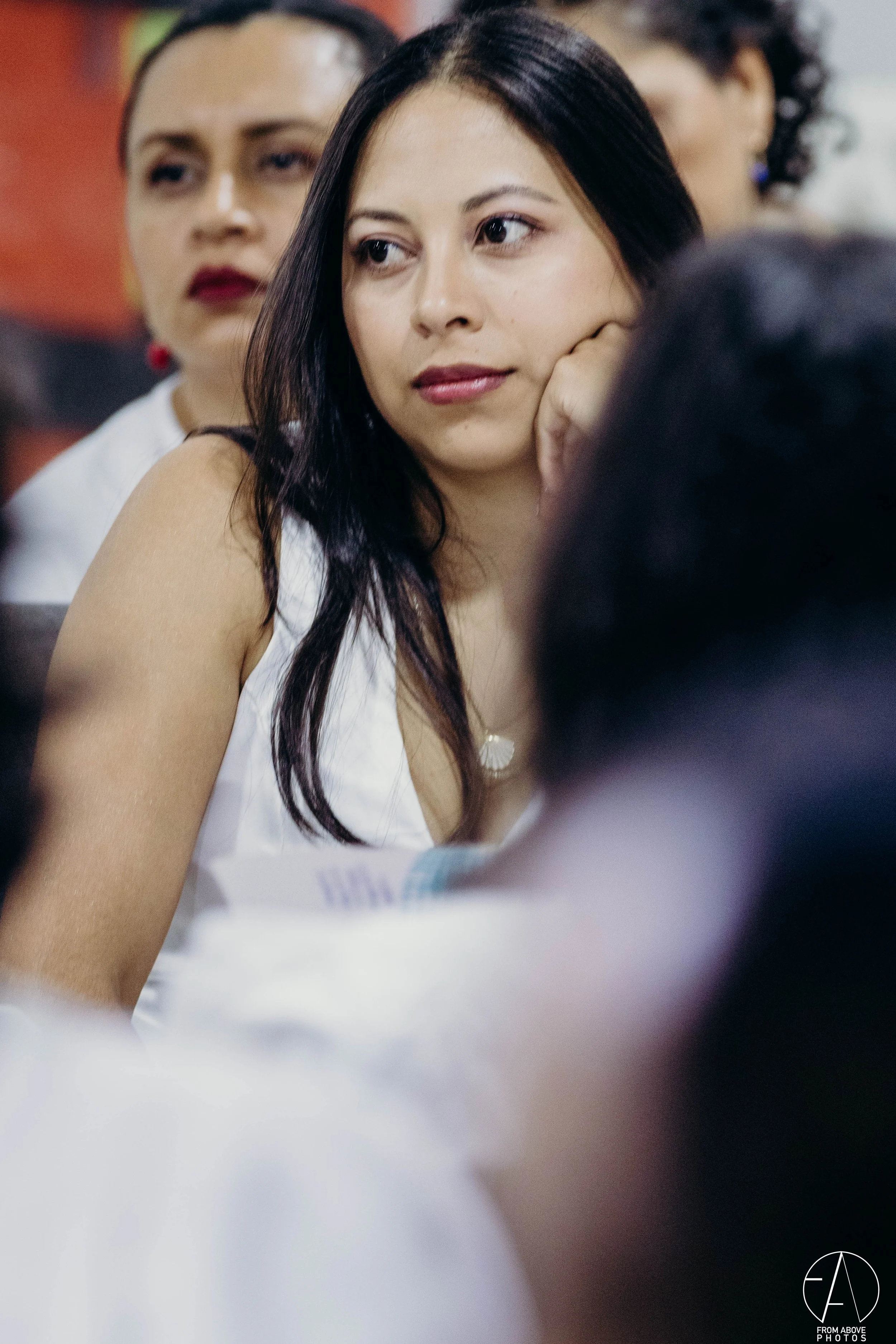 Mujer con cabello largo y oscuro, descansando su cabeza en su mano, en un entorno de varias personas en el fondo.