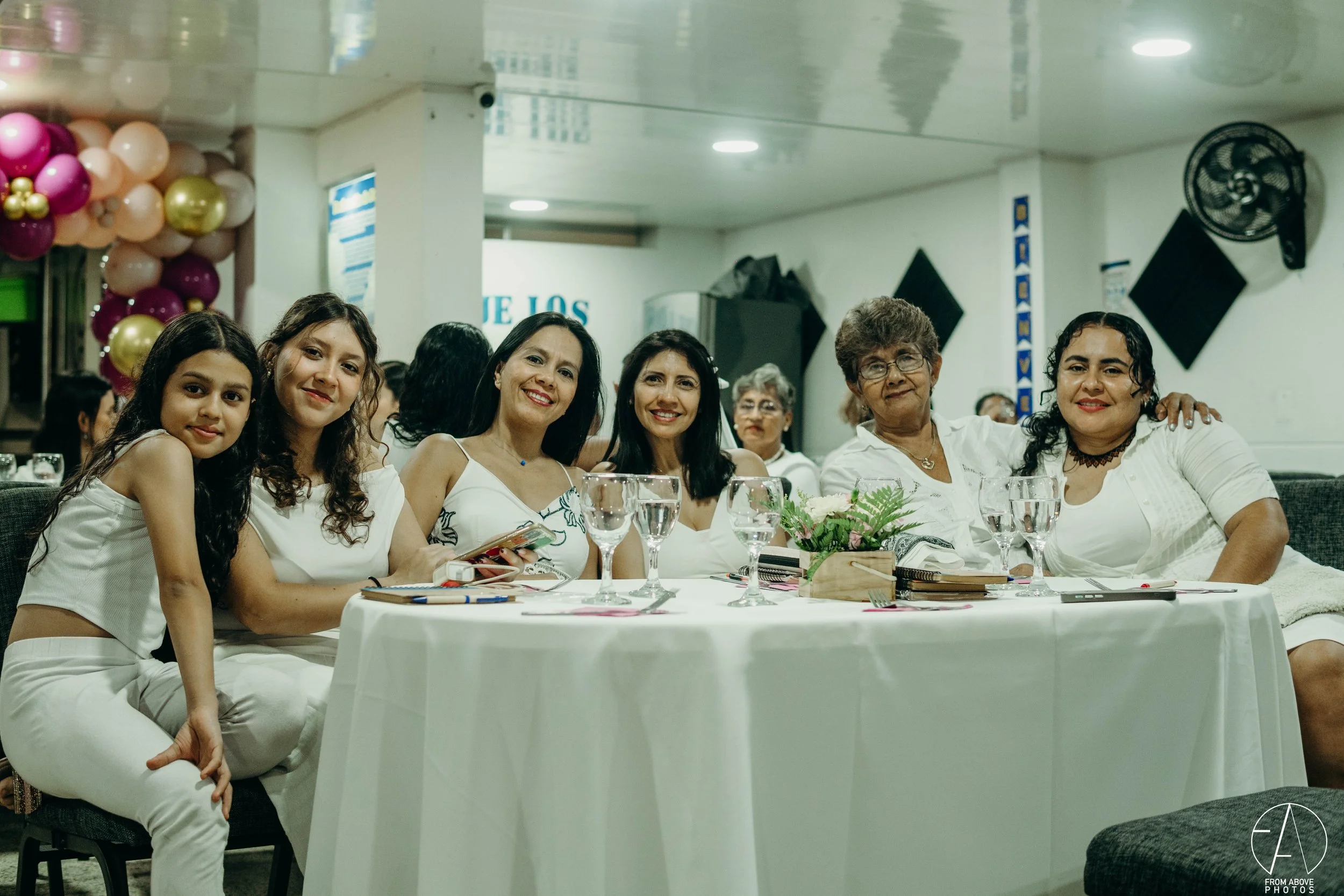 Grupo de siete mujeres sentadas en una mesa decorada con flores en un evento social, todas vestidas con ropa blanca, sonriendo y disfrutando la ocasión.