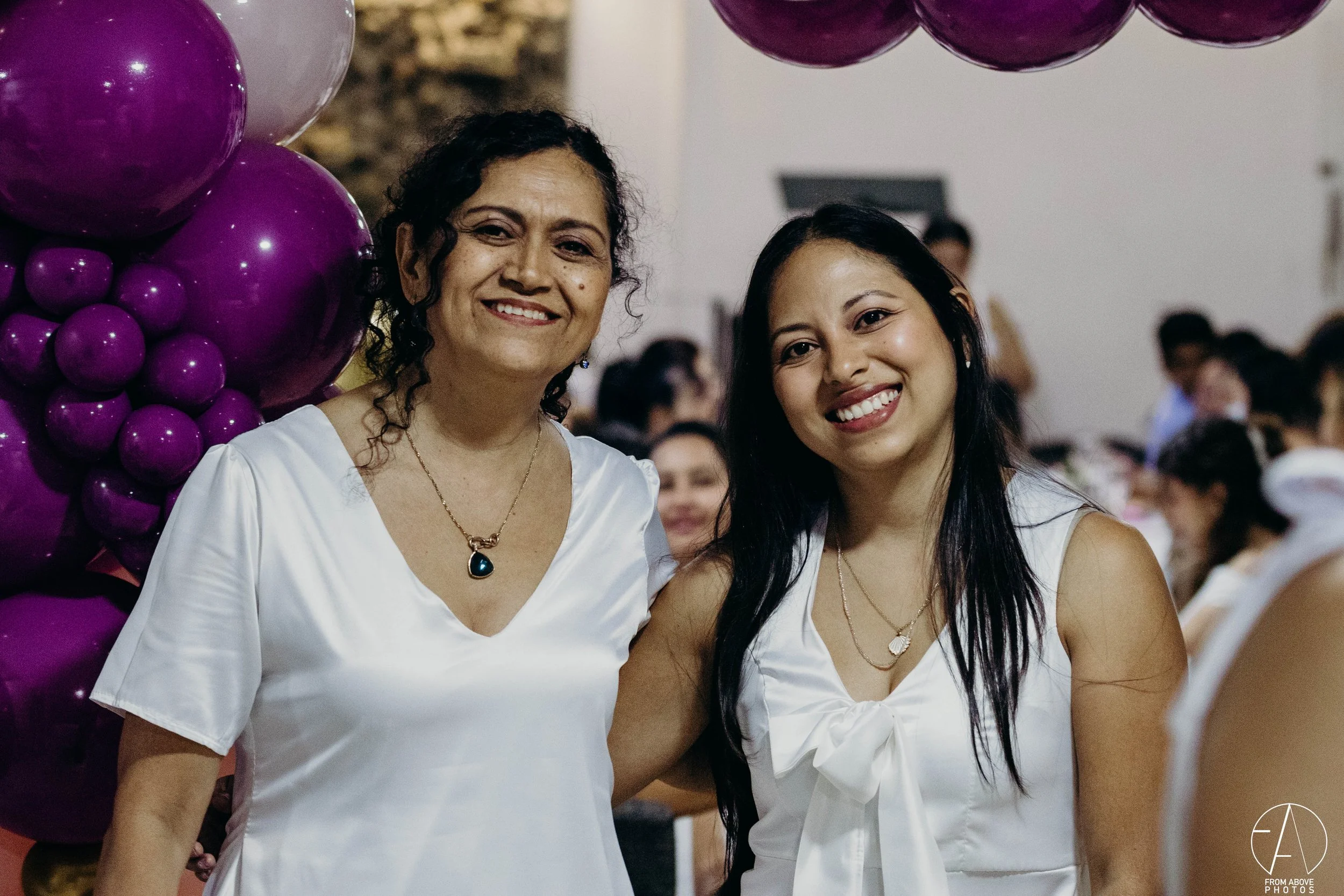 Dos mujeres sonriendo en un evento, con decoraciones de globos morados y personas en el fondo.