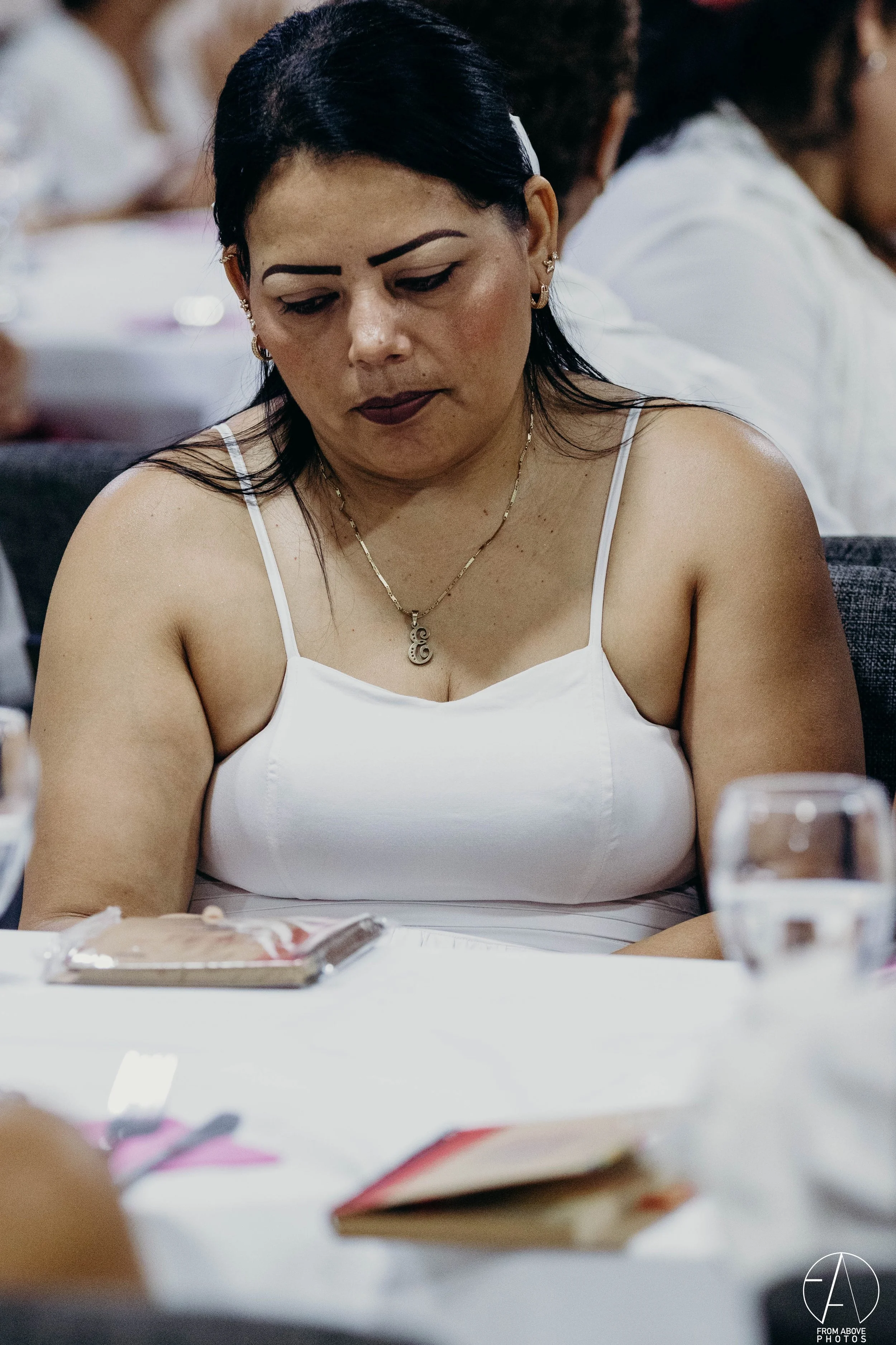 Mujer con cabello negro, piel clara, usando blusa blanca de tirantes y un collar dorado, concentrada en un libro en una mesa de comedor, en un entorno de reunión social.