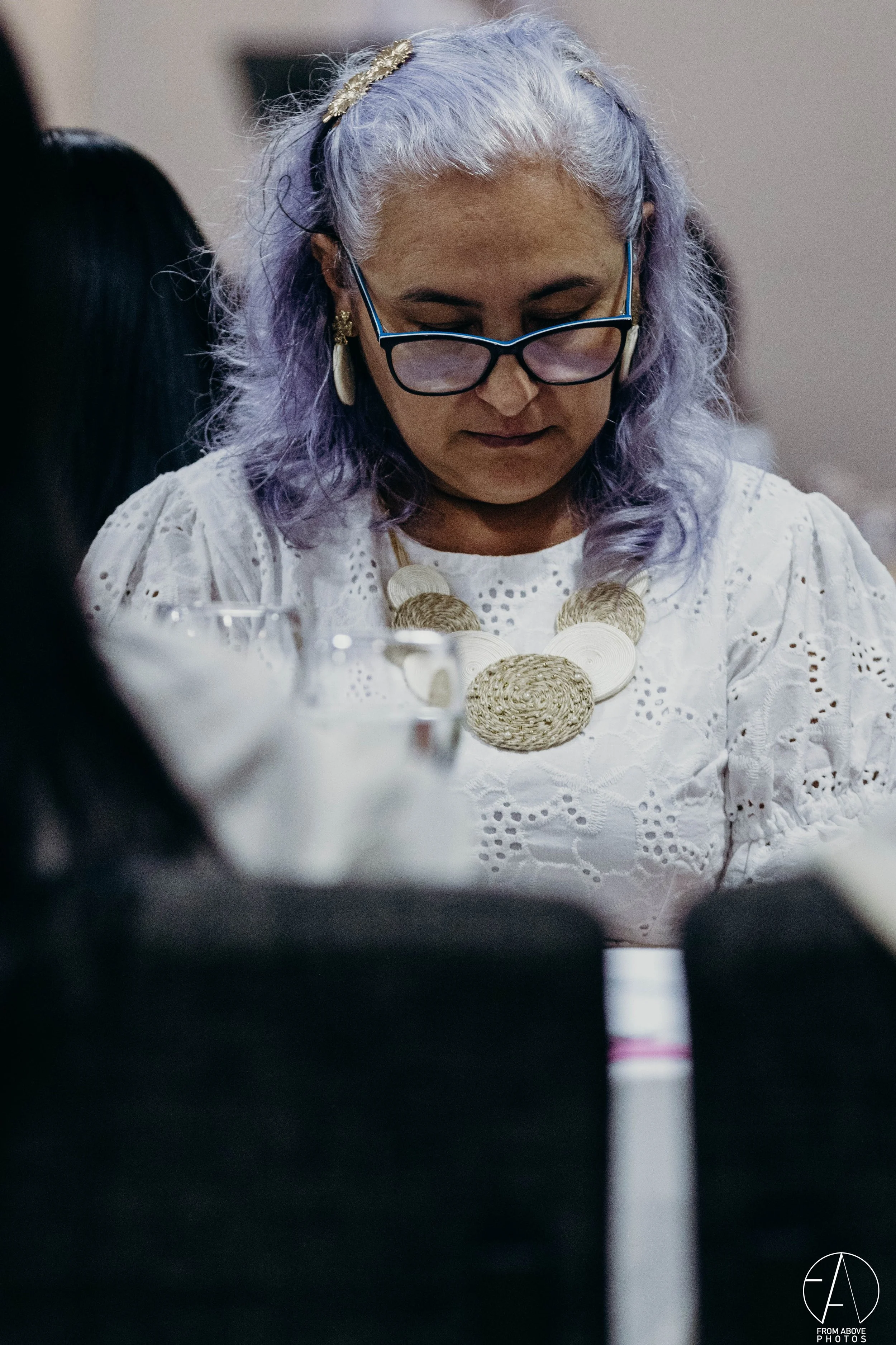 Mujer con cabello rubio y lila, usando gafas y un collar de cuerda, vista de frente, en un entorno interior con otras personas alrededor.