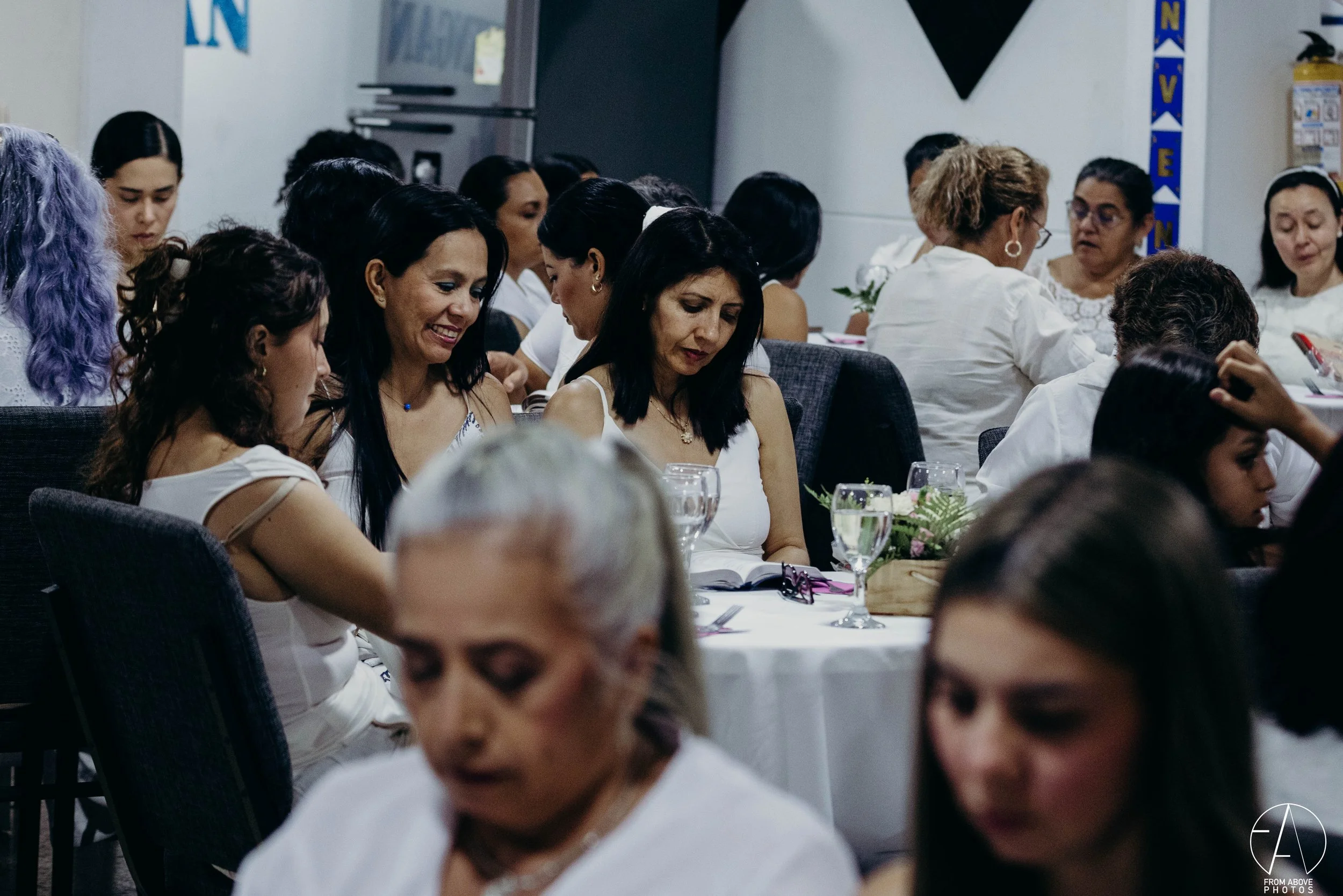 Un grupo de mujeres sentadas en una mesa, algunas con sus ojos cerrados o mirando hacia abajo, participando en una actividad espiritual o de oración. Todas llevan ropa blanca y parecen estar en un ambiente de reunión o evento comunitario.