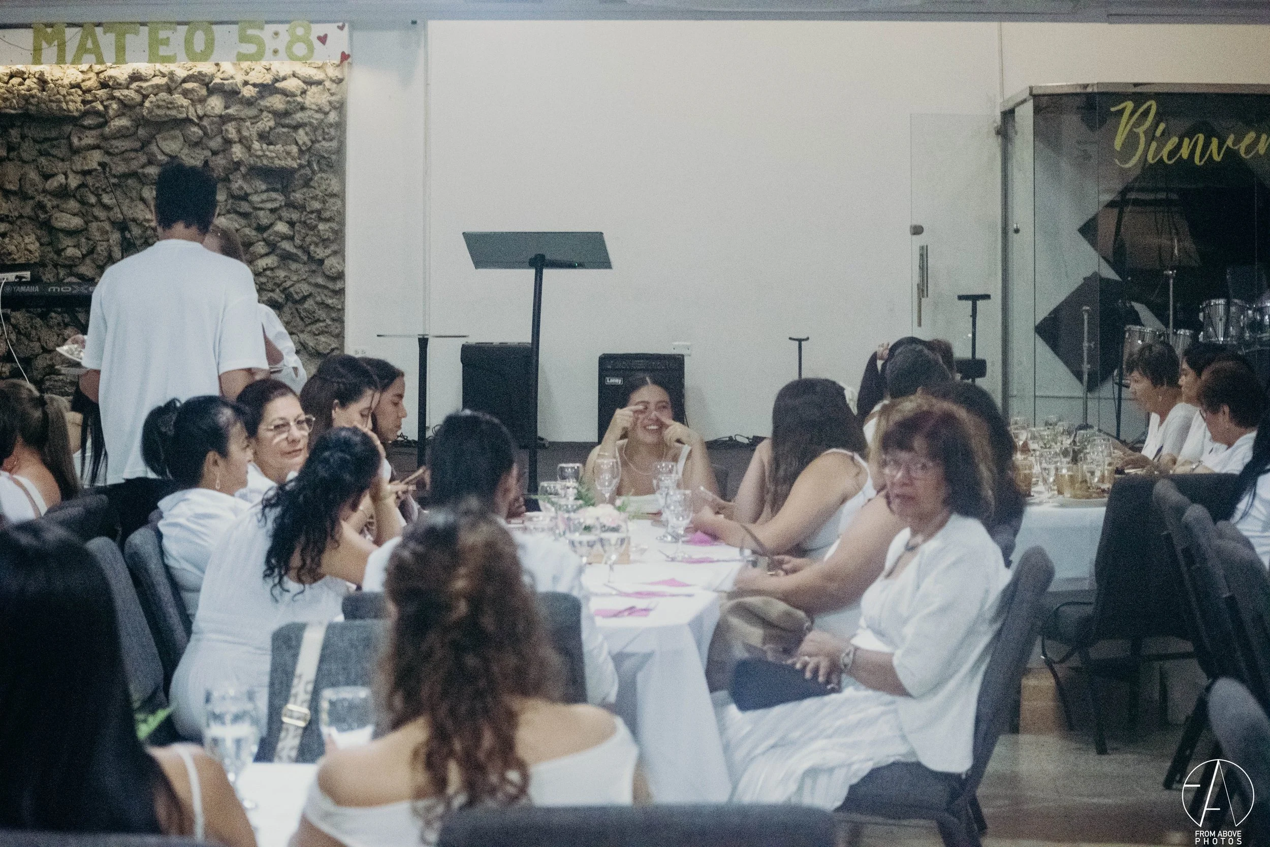 Grupo de personas sentadas alrededor de una mesa en un evento, algunas de ellas riendo y hablando, en un ambiente interior con decoraciones simples y equipo de sonido al fondo.